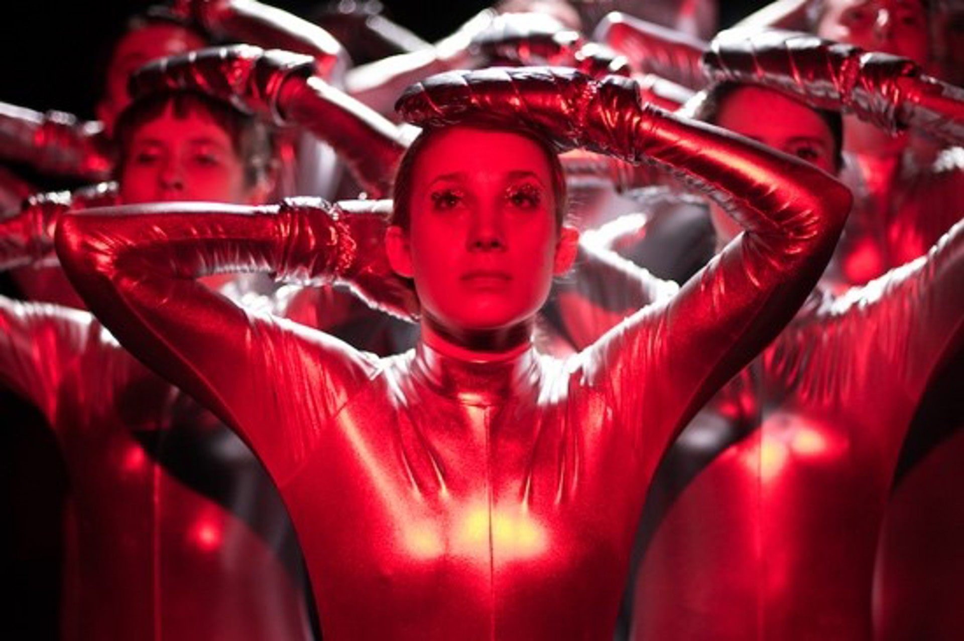 female with hands on head with other dancers in the background copying the movement. All wearing lycra bodysuit with red lighting. female with hands on head with other dancers in the background copying the movement. All wearing lycra bodysuit with red lighting.