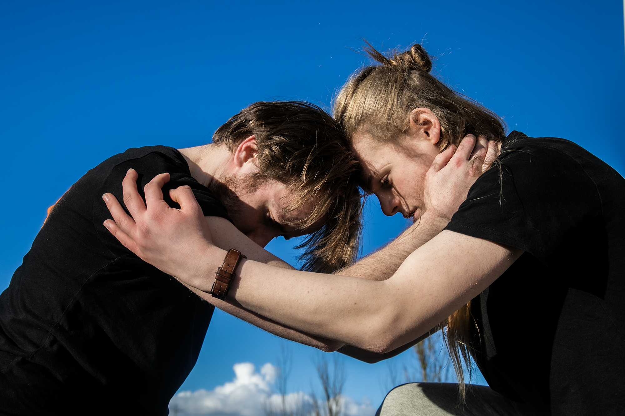 Two white male dancers holding eachothers forearms with heads together looking down with blue sky background. Two white male dancers holding eachothers forearms with heads together looking down with blue sky background.