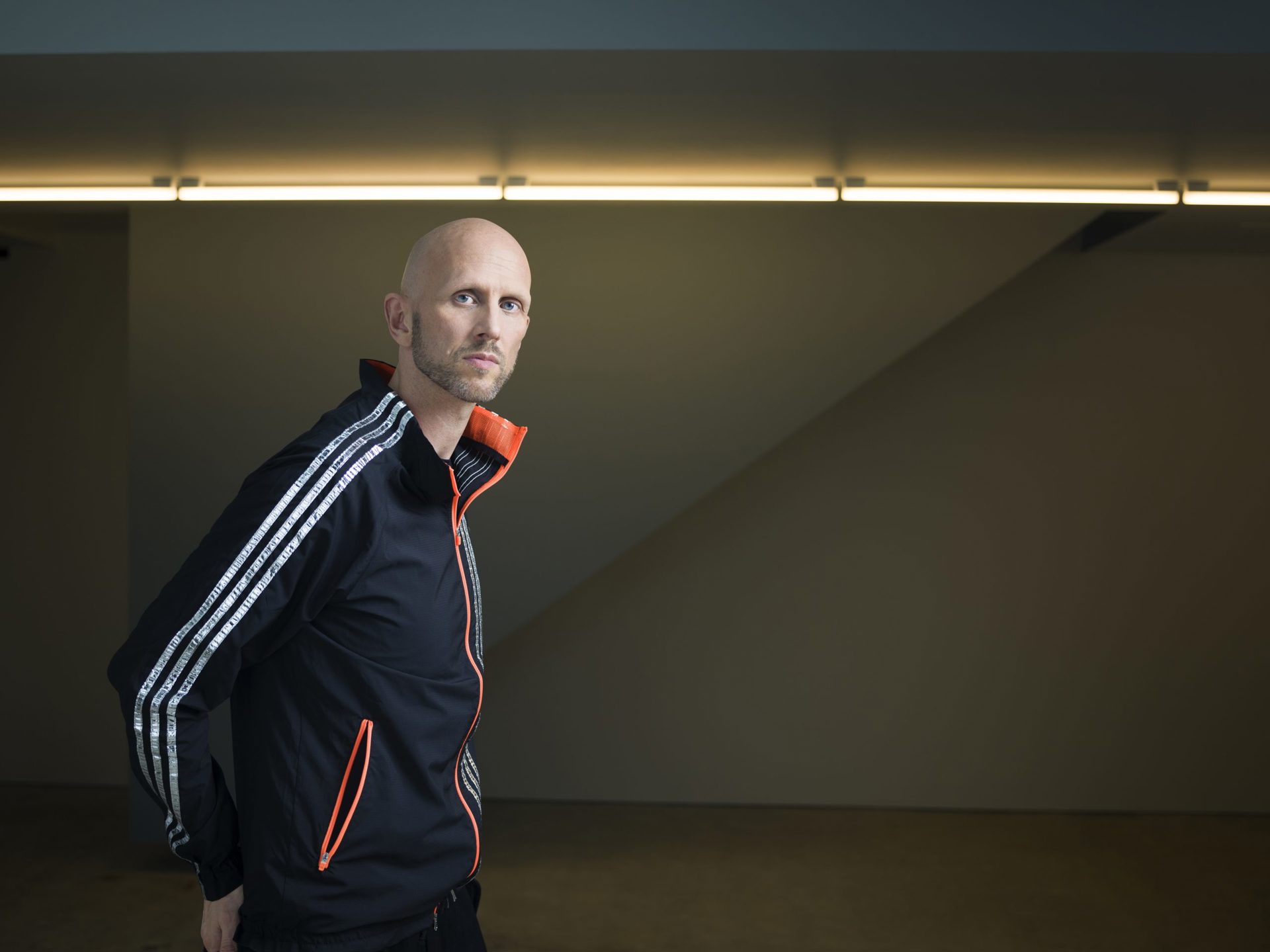headshot of Wayne Mcgregor. White bald male with stubble standing to the left of image wearing a blue tracksuit zip up 