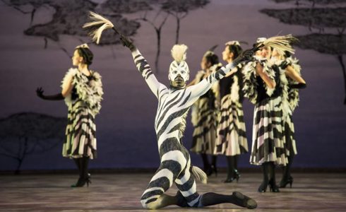 dancers on stage, one central dancer dressed as a zebra kneeled on the floor with arms stretched wide looking at the camera. The backdrop of the southern cape with trees.