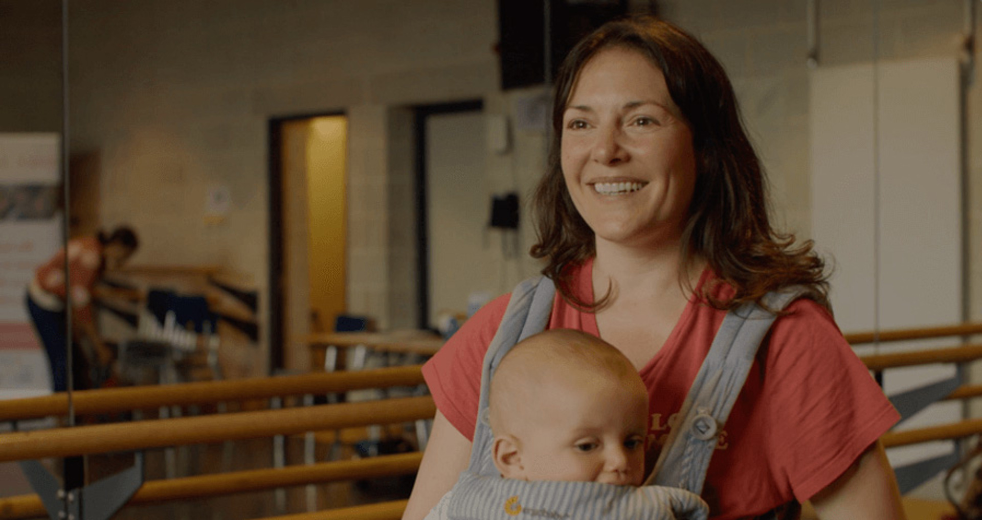 White female holding baby in a body carrier talking to the camera in a dance studio White female holding baby in a body carrier talking to the camera in a dance studio