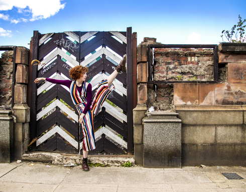 white female dancer with red short curly hair and striped jumpsuit, high kicking right leg with left hand to the side. In front of triangle painted black and white gate with brick wall and blue sky above. white female dancer with red short curly hair and striped jumpsuit, high kicking right leg with left hand to the side. In front of triangle painted black and white gate with brick wall and blue sky above.
