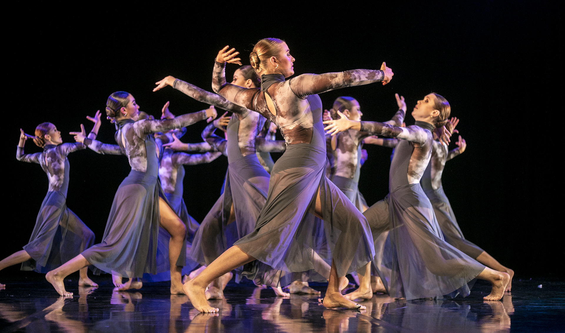 Severn female dancers on a stage wearing long purple jumpsuits with legs in a stride, arched back and arms in front of them scattered around the stage.
