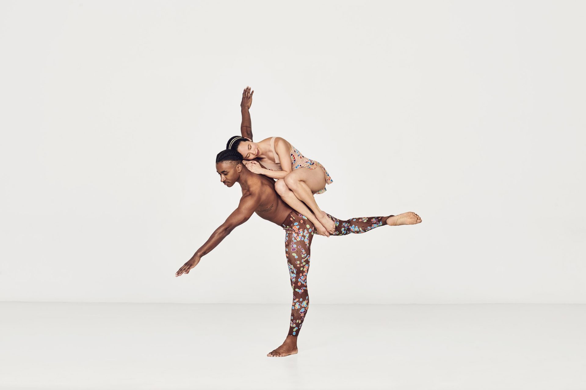 Global majority male dancer on one leg with arms stretched wise with global majority female dancer curled up balancing in his back in a white studio Global majority male dancer on one leg with arms stretched wise with global majority female dancer curled up balancing in his back in a white studio