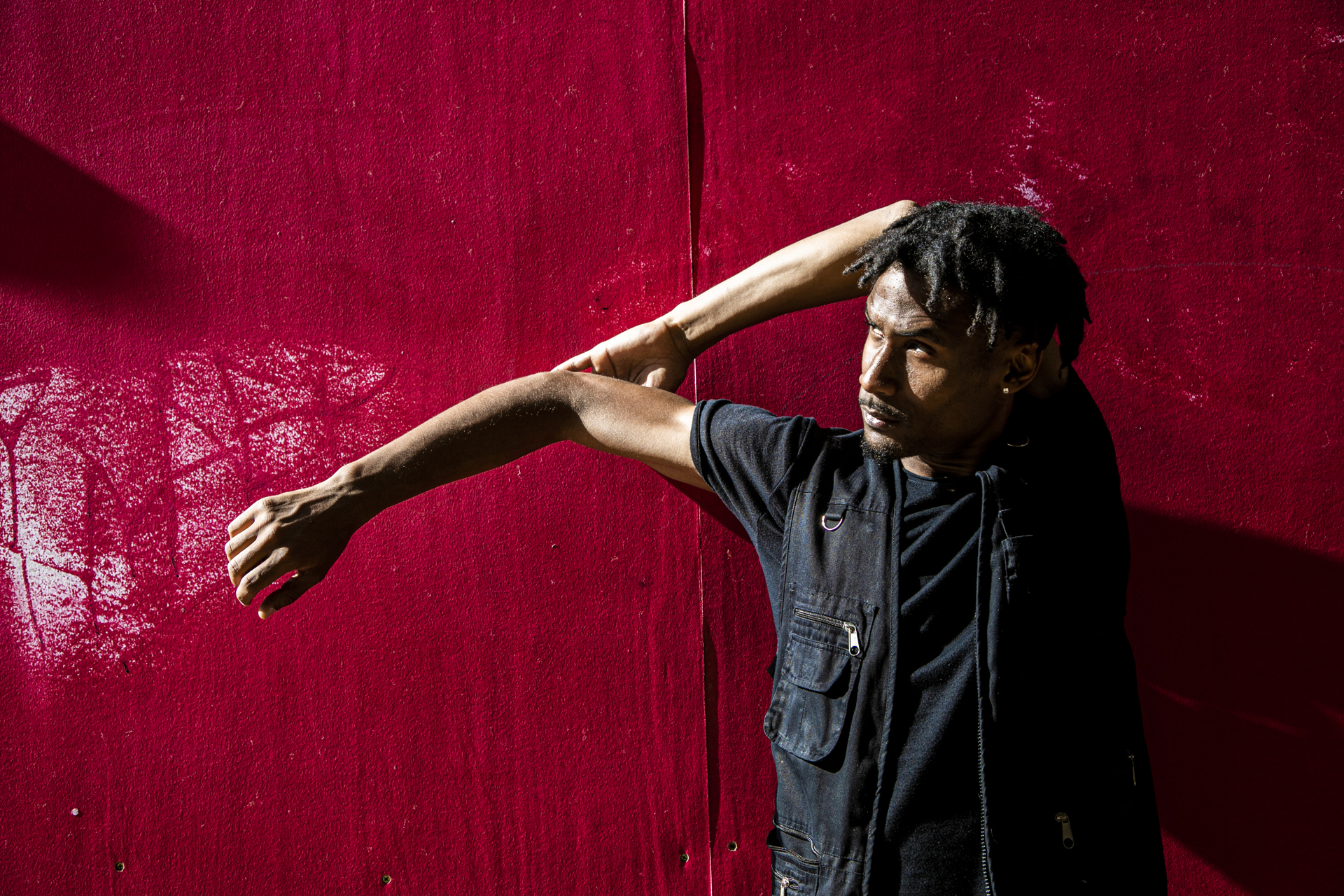 Global majority male dancer with short dreaded hair, one arm strecthed to the left and the other holding the elbow behind his head. Wearing all black standing in front of a red wall . Global majority male dancer with short dreaded hair, one arm strecthed to the left and the other holding the elbow behind his head. Wearing all black standing in front of a red wall .