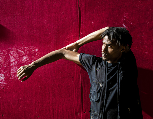 Global majority male dancer with short dreaded hair, one arm strecthed to the left and the other holding the elbow behind his head. Wearing all black standing in front of a red wall . Global majority male dancer with short dreaded hair, one arm strecthed to the left and the other holding the elbow behind his head. Wearing all black standing in front of a red wall .