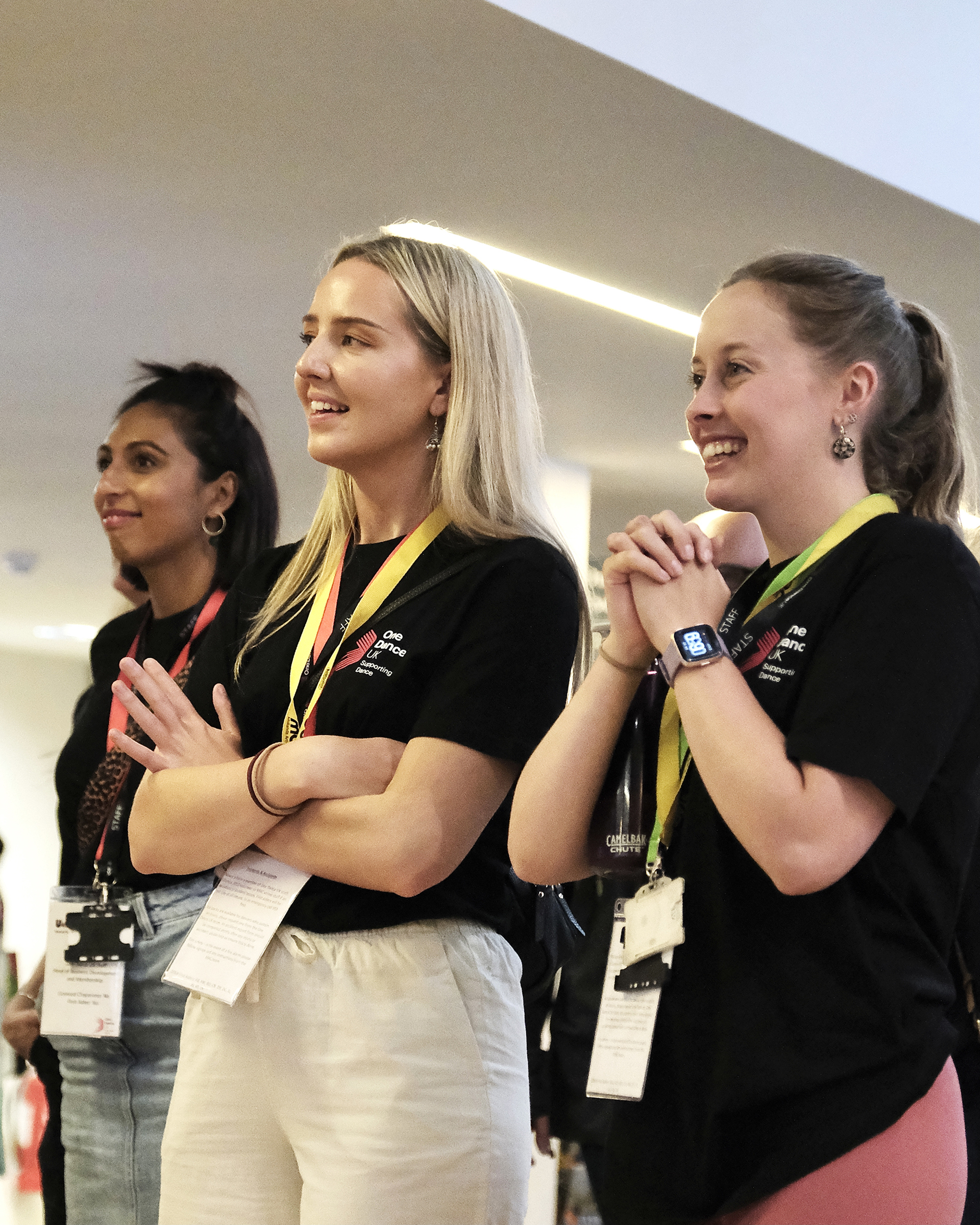 Three female One Dance UK staff members smiling in a foyer whilst watching a performance Three female One Dance UK staff members smiling in a foyer whilst watching a performance