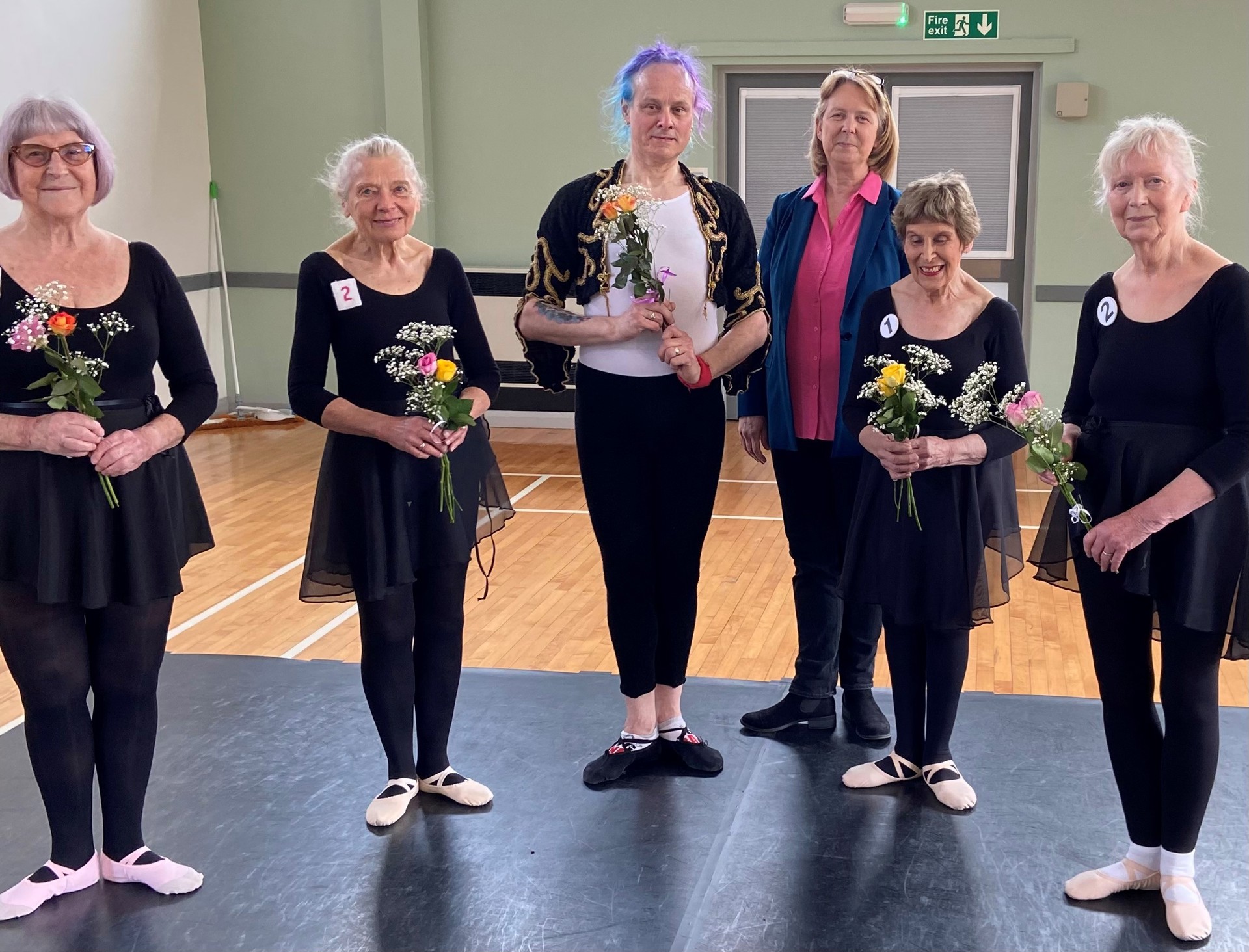 Six older white Bbodance dancers wearing black ballet costumes holding flowers and looking at the camera. In a dance studio, Six older white Bbodance dancers wearing black ballet costumes holding flowers and looking at the camera. In a dance studio,