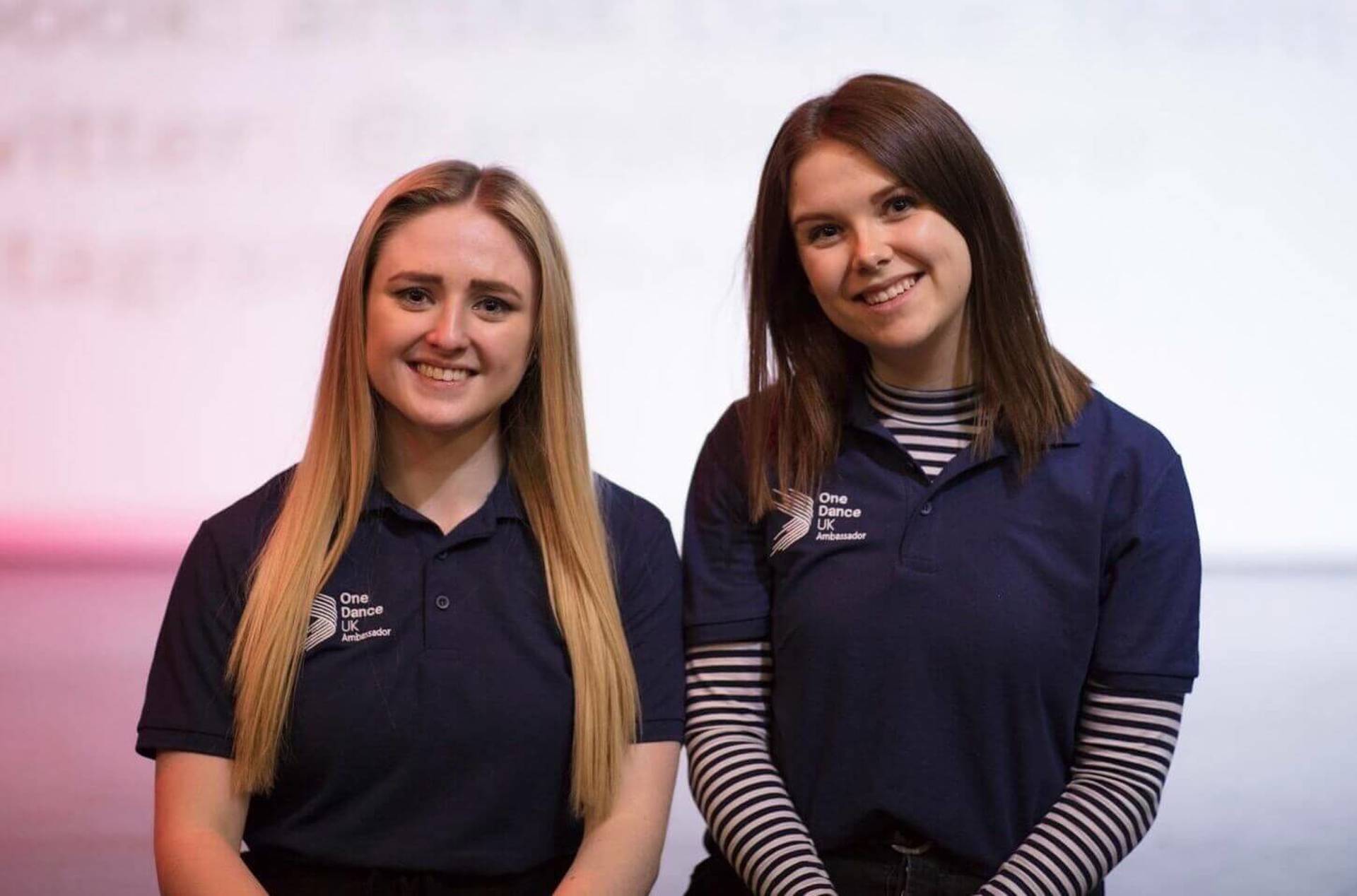 Two female One Dance UK dance ambassadors in blue t-shirts smiling at the camera Two female One Dance UK dance ambassadors in blue t-shirts smiling at the camera