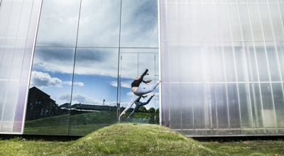 female dancer jumping with one leg pointed towards the ground and the other kicked behind witharms in the air on a grassey hill. In front of metal Trinity Laban University building  with mirroed wall reflecting clouded sky. Wearing grey leggings and black crop top.  female dancer jumping with one leg pointed towards the ground and the other kicked behind witharms in the air on a grassey hill. In front of metal Trinity Laban University building  with mirroed wall reflecting clouded sky. Wearing grey leggings and black crop top.