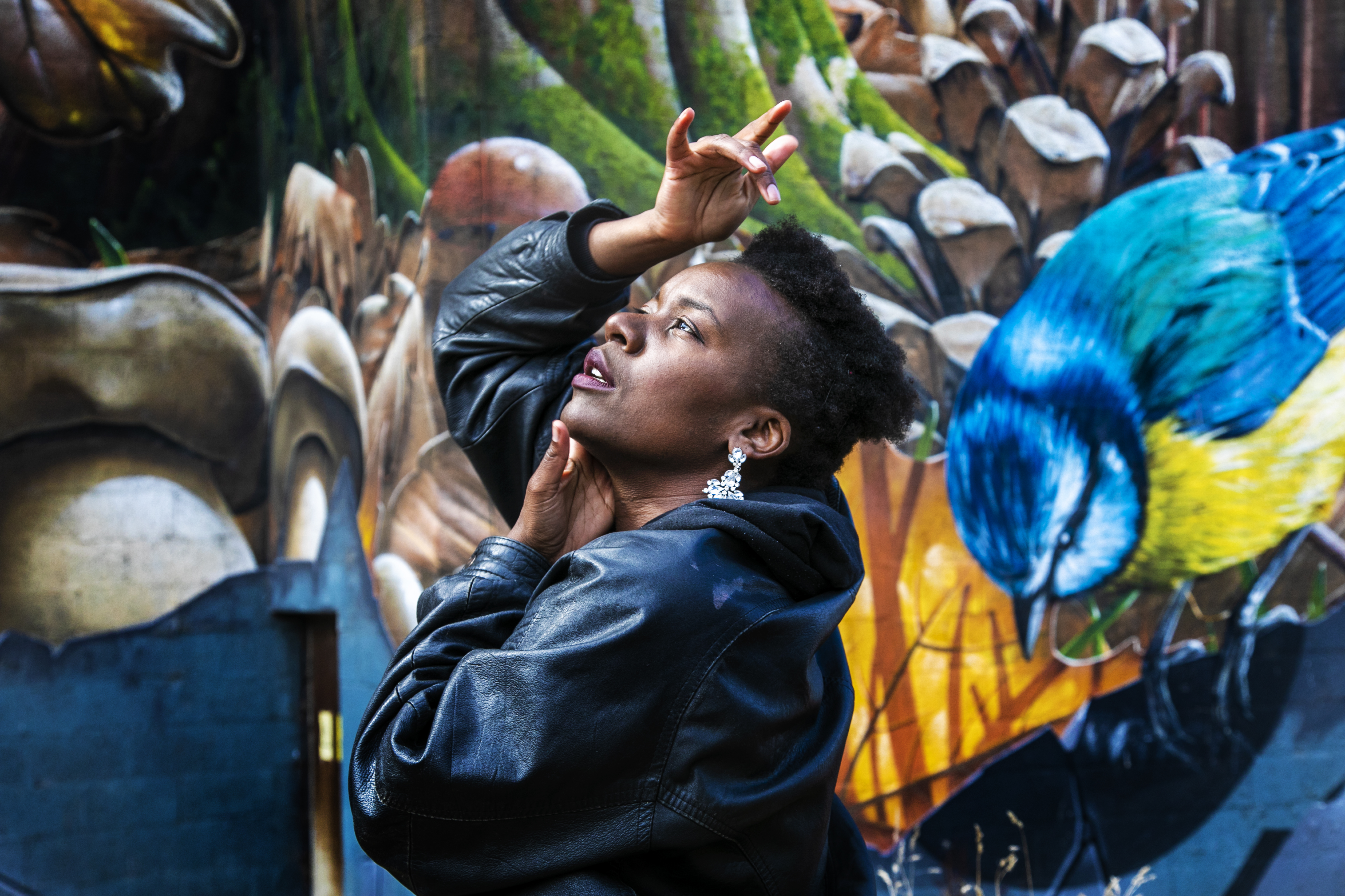Global Majority female looking up to the top left of the image with hands around her face. Wearing a black leather jacket with short afro hair. In front of colourful graffiti wall Global Majority female looking up to the top left of the image with hands around her face. Wearing a black leather jacket with short afro hair. In front of colourful graffiti wall