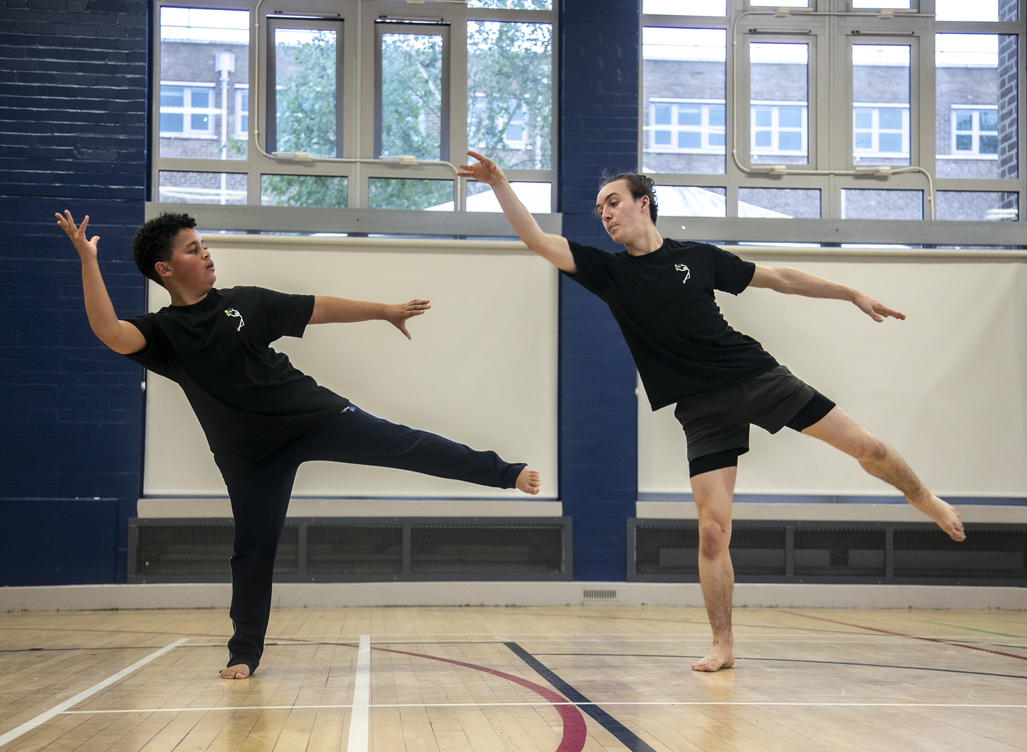 teenage male dance teacher on one leg reaching to the top left of the image teaching young boy dancer who is mirroring the arms and the legs. In sports hall both wearing black PE kit