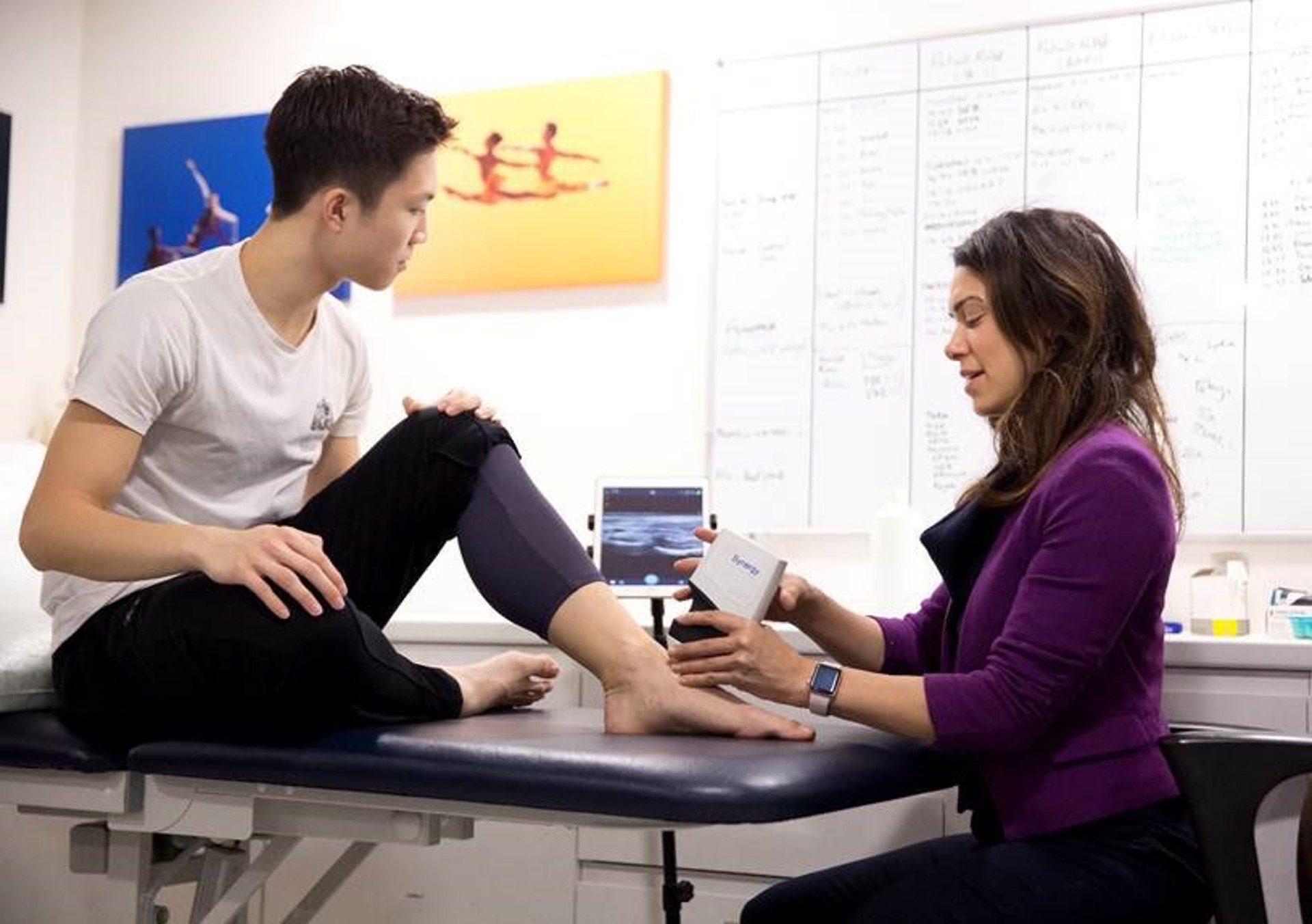 Young male dancer sitting on a physio bed with his foot out and a healthcare practitioner ultra sounding ankle with screen behind