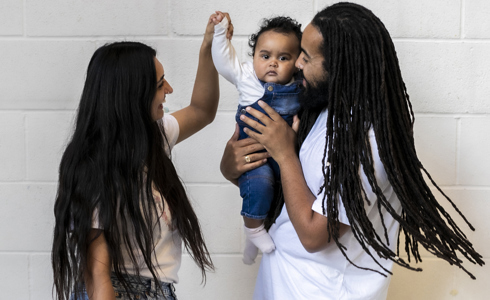 Family of 3. Global majority male with long dreadlocked hair holding baby, global majority female with long black hair, holding the babies hand as they dance. Wearing blue jeans, trainers and white tshirts infront of white brick wall. 