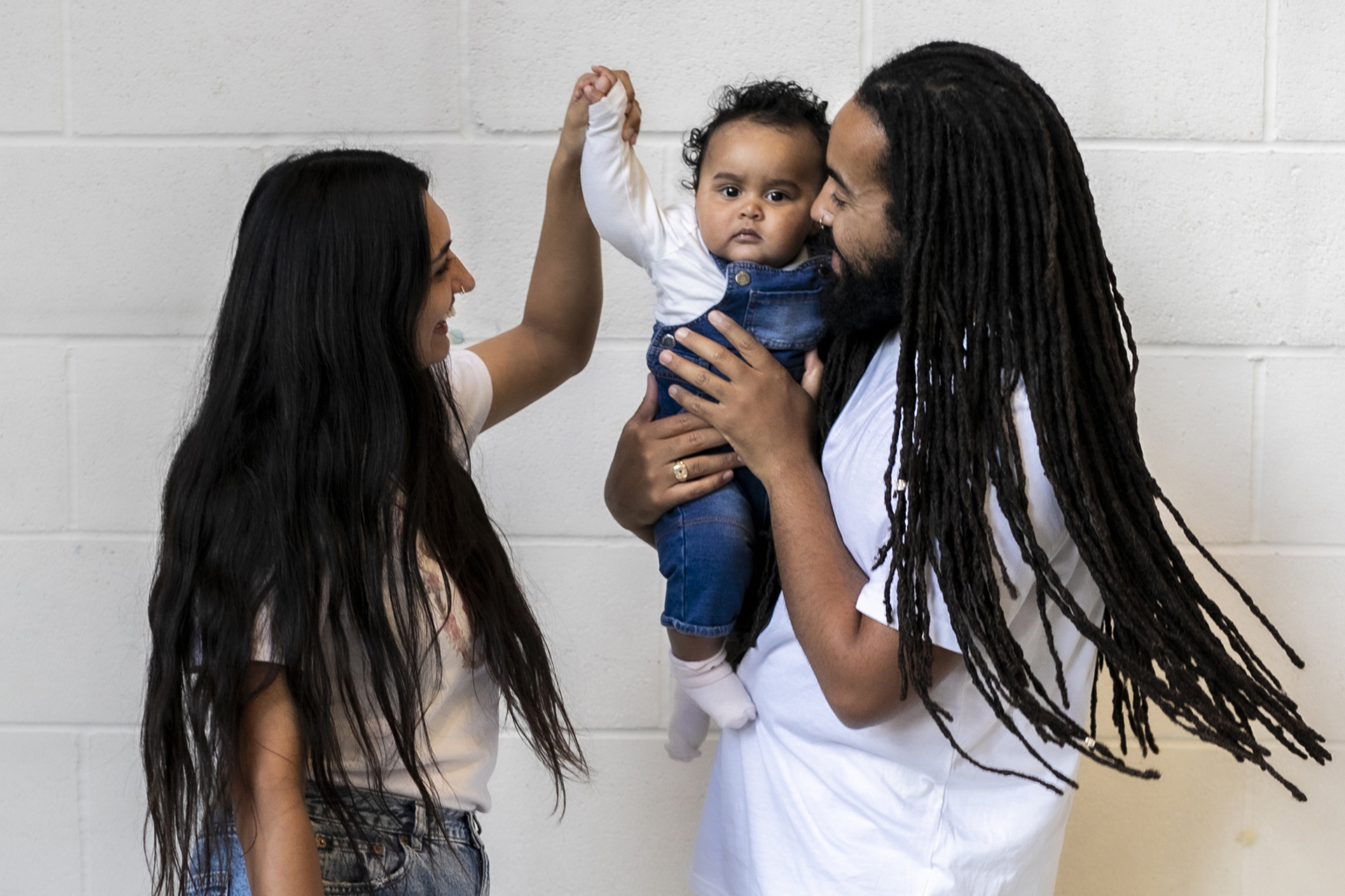 Family of 3. Global majority male with long dreadlocked hair holding baby, global majority female with long black hair, holding the babies hand as they dance. Wearing blue jeans, trainers and white tshirts infront of white brick wall. Family of 3. Global majority male with long dreadlocked hair holding baby, global majority female with long black hair, holding the babies hand as they dance. Wearing blue jeans, trainers and white tshirts infront of white brick wall.