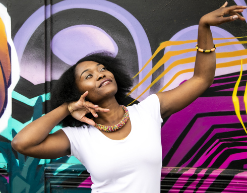 Global majority female dancer with afro, holding one arm out to the top right of the image with eyes looking at a limp wrist and the other hand next to her face. Wearing white top and colourful beaded necklace in fornt of coloured graffiti wall