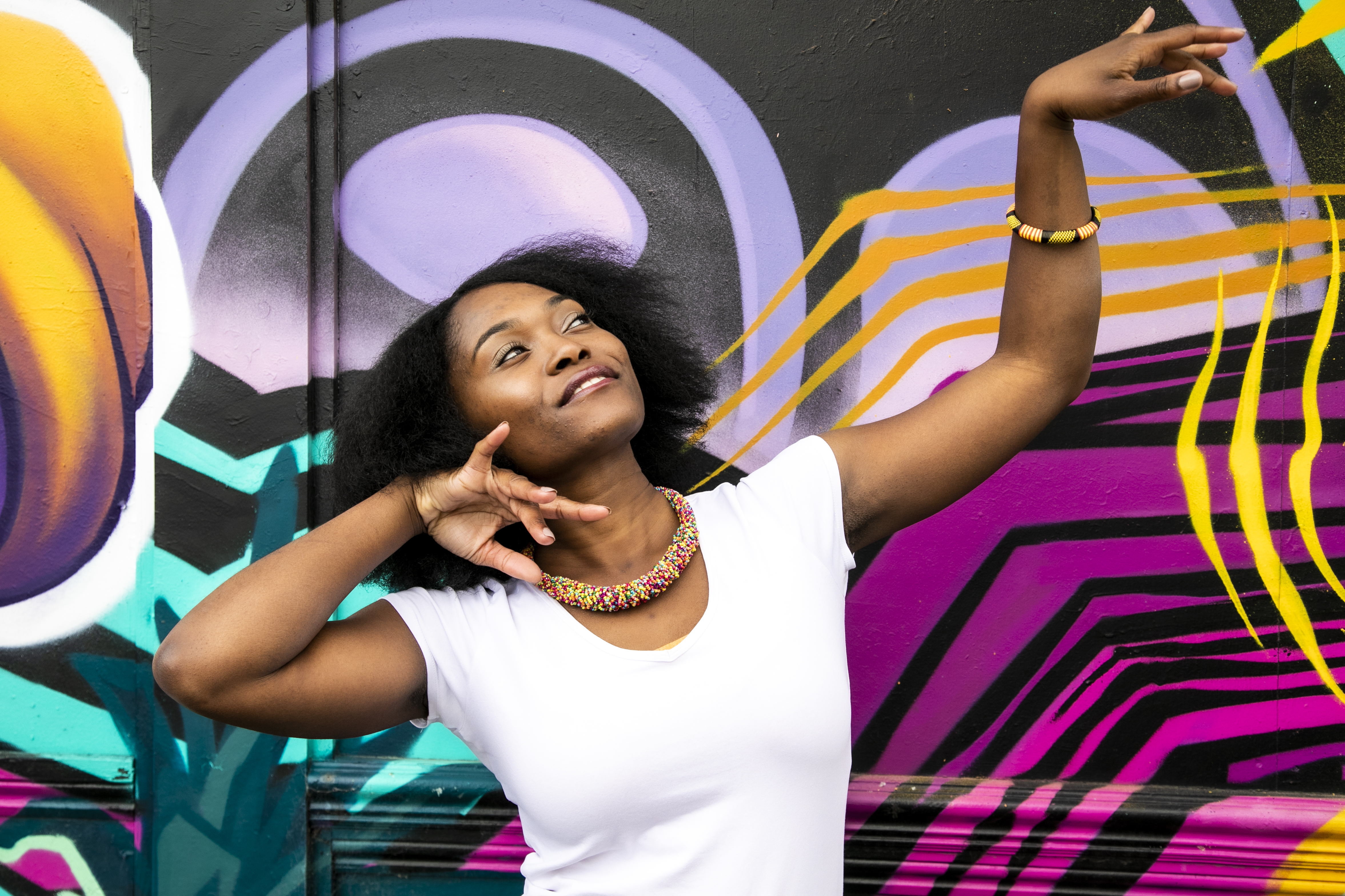 Global majority female dancer with afro, holding one arm out to the top right of the image with eyes looking at a limp wrist and the other hand next to her face. Wearing white top and colourful beaded necklace in fornt of coloured graffiti wall Global majority female dancer with afro, holding one arm out to the top right of the image with eyes looking at a limp wrist and the other hand next to her face. Wearing white top and colourful beaded necklace in fornt of coloured graffiti wall