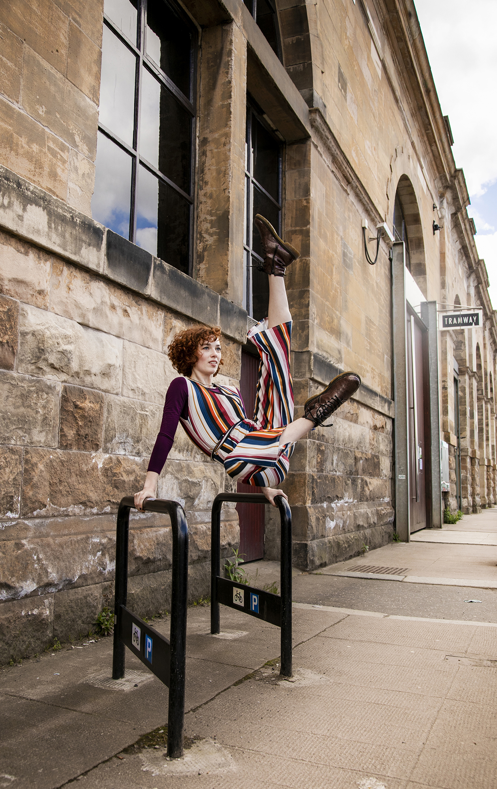 white female dancer with red short curly hair and striped jumpsuit, swinging on two metal bike stands high kicking both legs in the air. In front tramway brick building on the street. white female dancer with red short curly hair and striped jumpsuit, swinging on two metal bike stands high kicking both legs in the air. In front tramway brick building on the street.