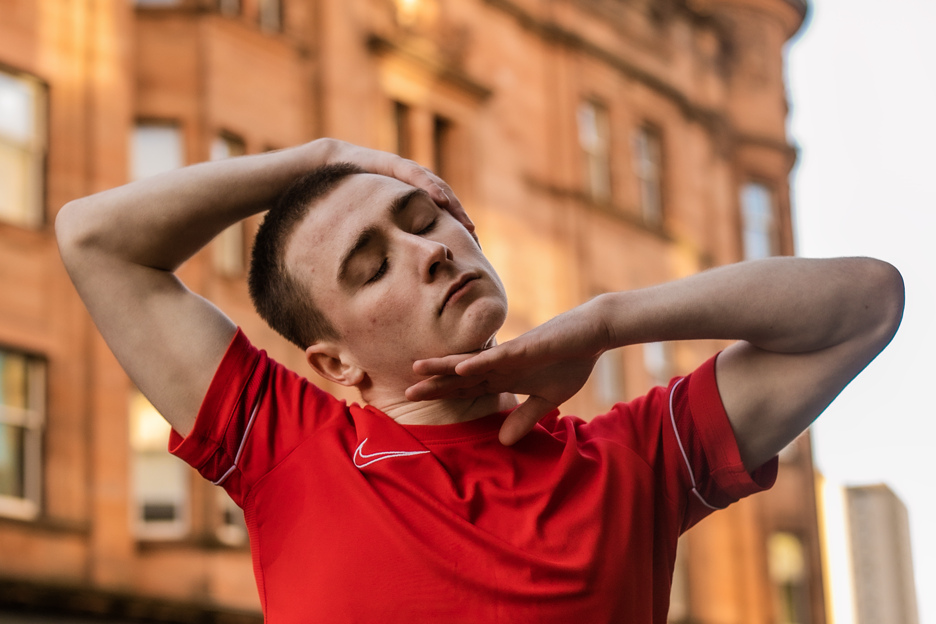 white male dancer with shaved head and red t shirt. On the street infront of orange building with hands around the face white male dancer with shaved head and red t shirt. On the street infront of orange building with hands around the face