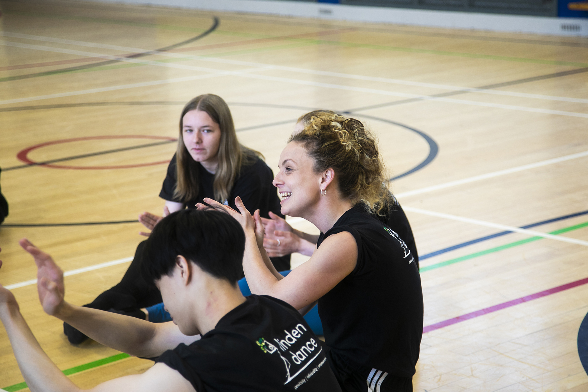 white female teacher with teenage student sitting on a sports hall floor with hands gesturing. All wearing black with linden dance logo on the t-shirts.  white female teacher with teenage student sitting on a sports hall floor with hands gesturing. All wearing black with linden dance logo on the t-shirts.