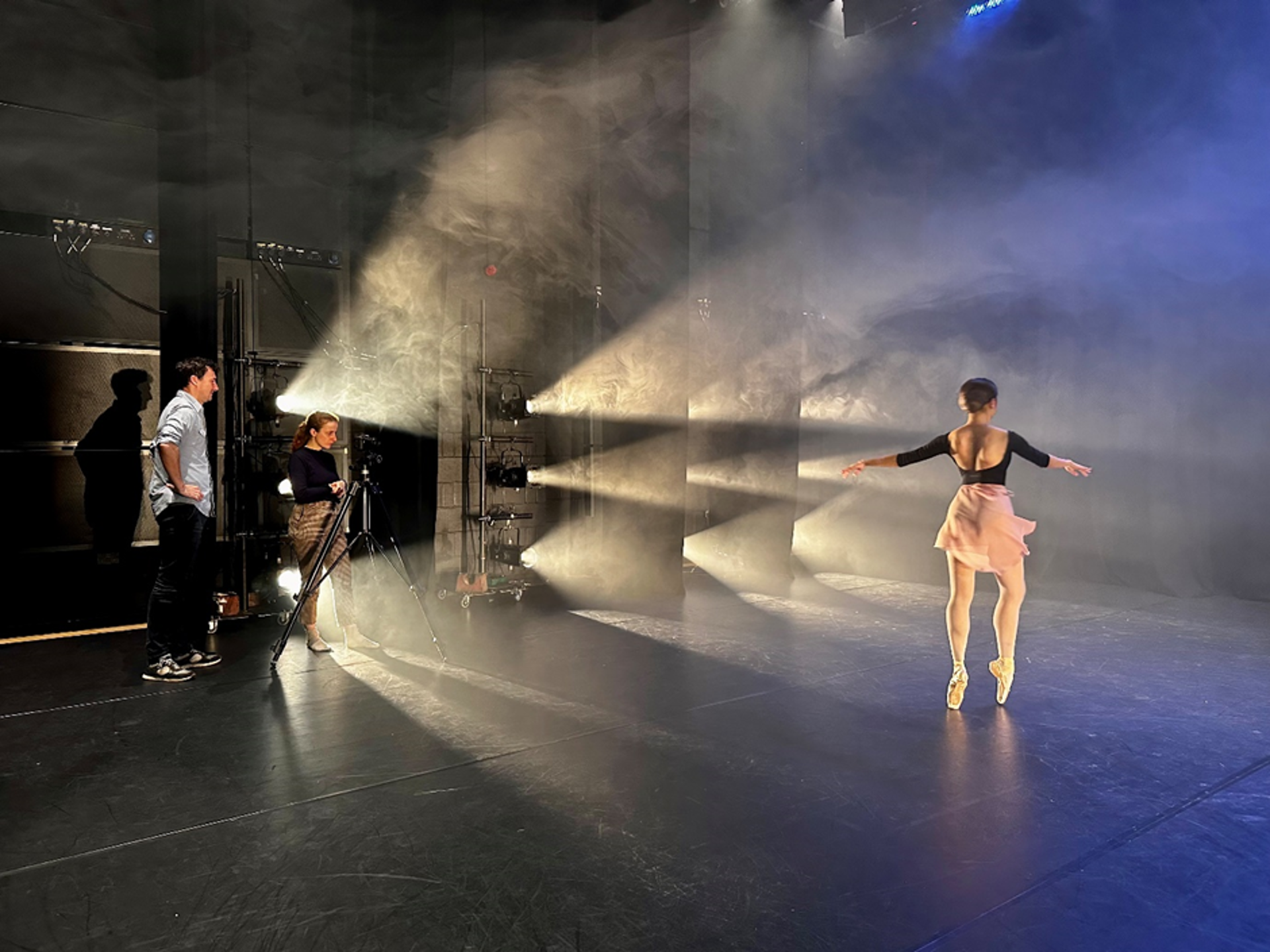 Female dancer on point on stage with stage lights facing her and a two people standing by a camera filming her