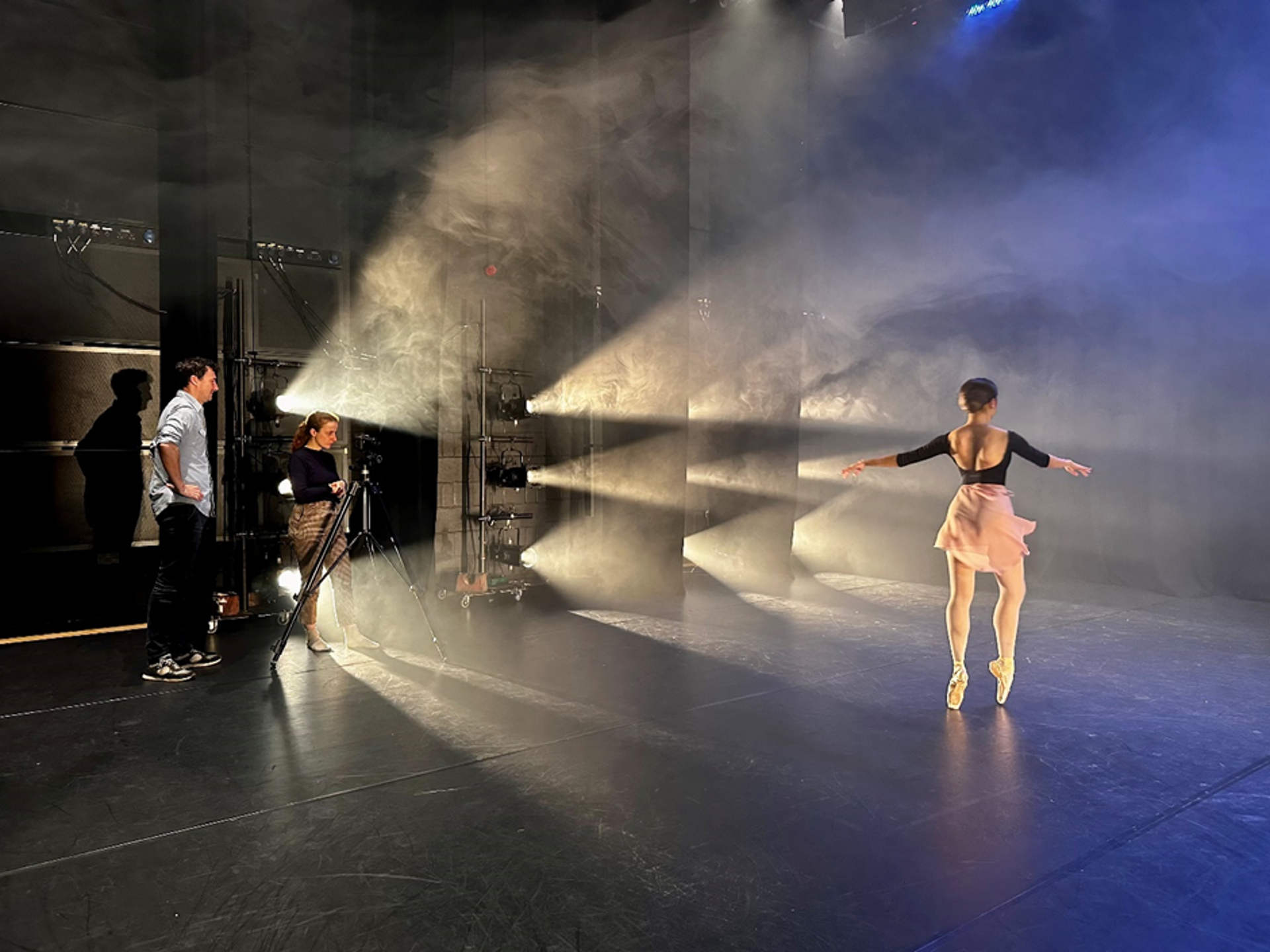 Female dancer on point on stage with stage lights facing her and a two people standing by a camera filming her Female dancer on point on stage with stage lights facing her and a two people standing by a camera filming her