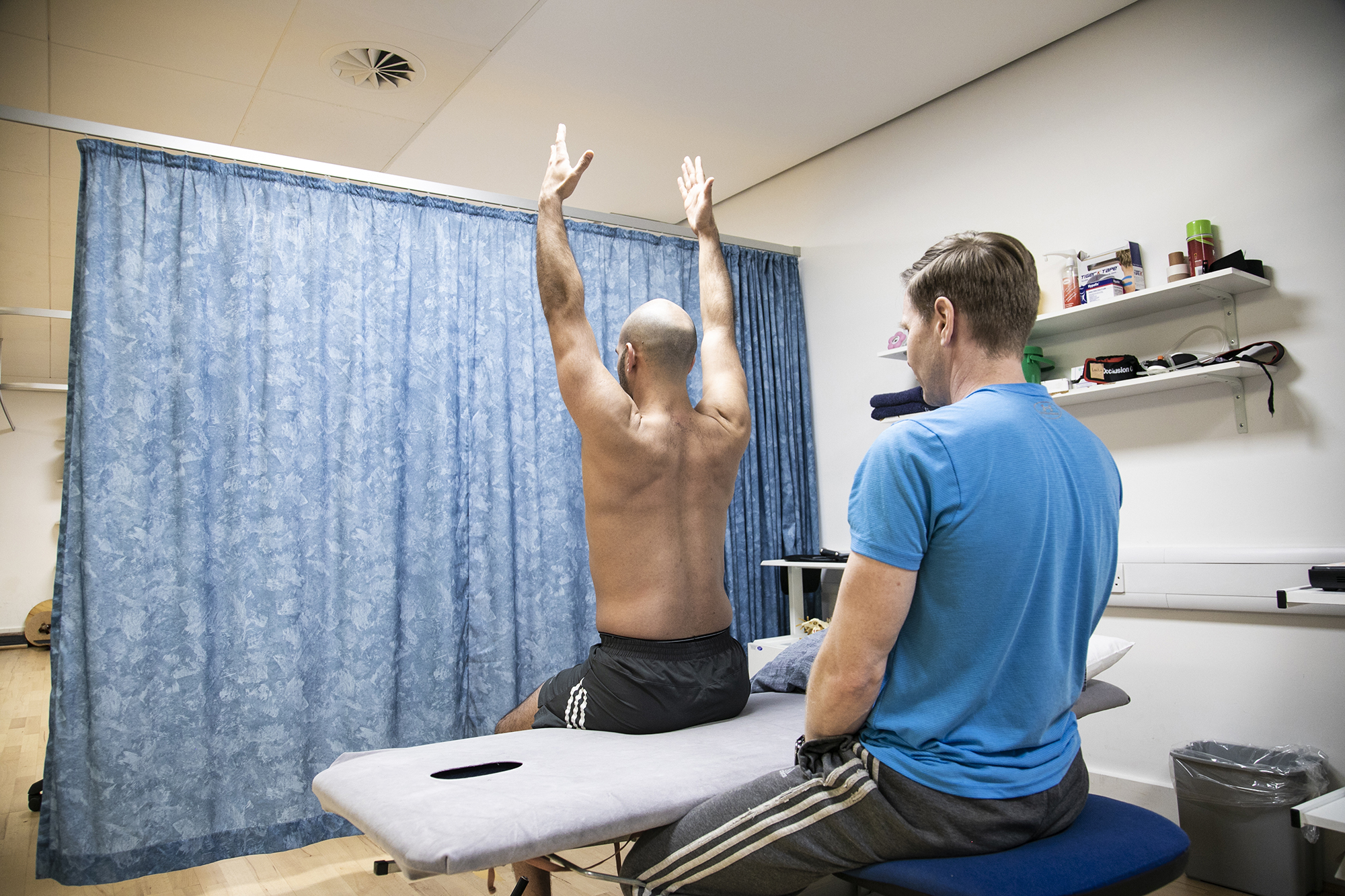 Male dancer sitting on physio bed with back to camera and arms up in the air, with male physiotherapist looking at dancer in an examination. With blue curtain in the background and a shelf of physio equipment.  Male dancer sitting on physio bed with back to camera and arms up in the air, with male physiotherapist looking at dancer in an examination. With blue curtain in the background and a shelf of physio equipment.
