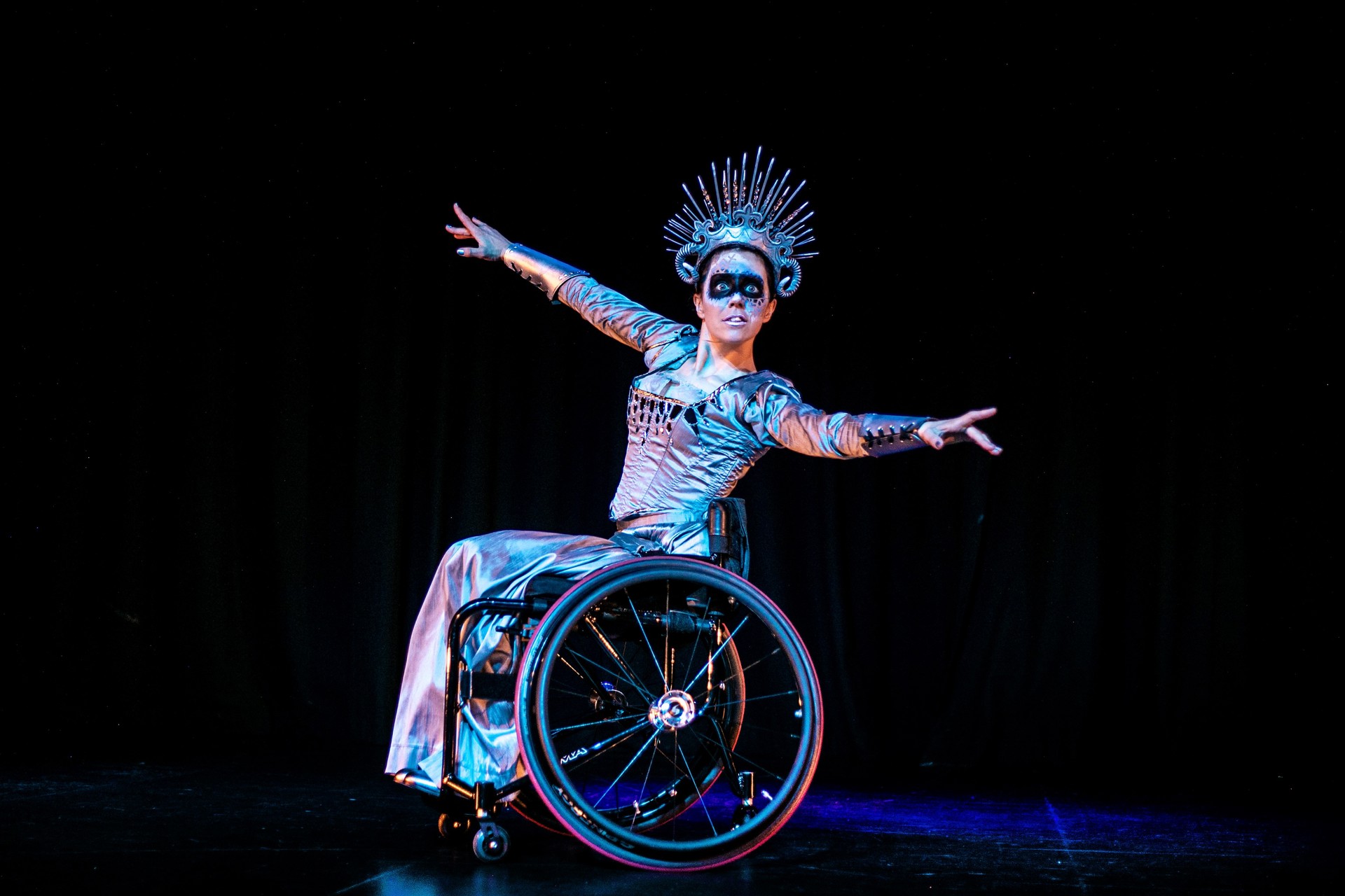 White female wheelchair dancer on stage wearing snow queen costume including large spike crown and black face paint across the eyes White female wheelchair dancer on stage wearing snow queen costume including large spike crown and black face paint across the eyes