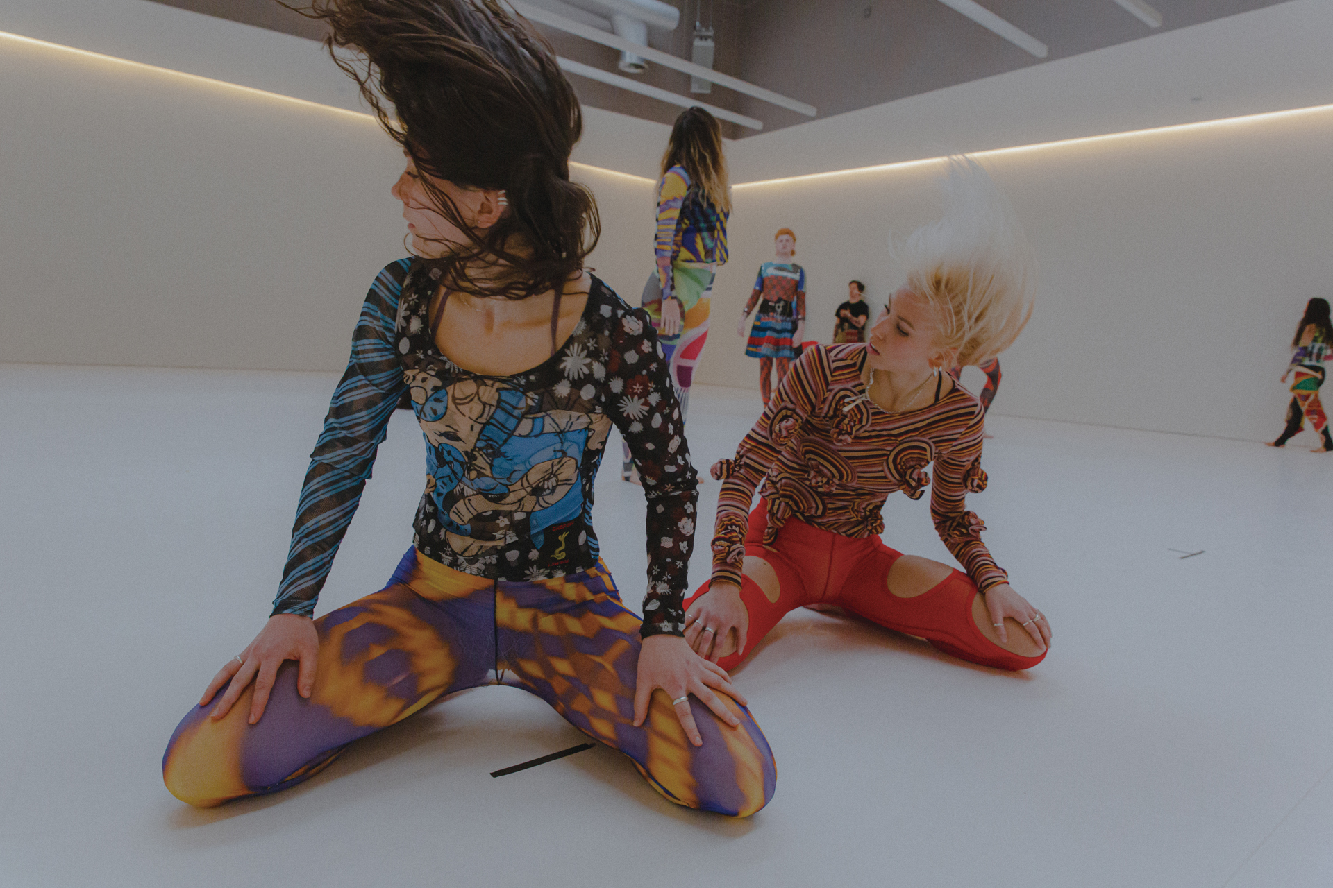 two female dancers sitting on their knees with hands on knees looking left and flicking hair. Wearing colourful patterned clothes two female dancers sitting on their knees with hands on knees looking left and flicking hair. Wearing colourful patterned clothes