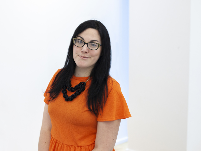 Headshot of Lara Coffey white female with black long hair and glasses smiling at the camera. Wearing orange top  Headshot of Lara Coffey white female with black long hair and glasses smiling at the camera. Wearing orange top