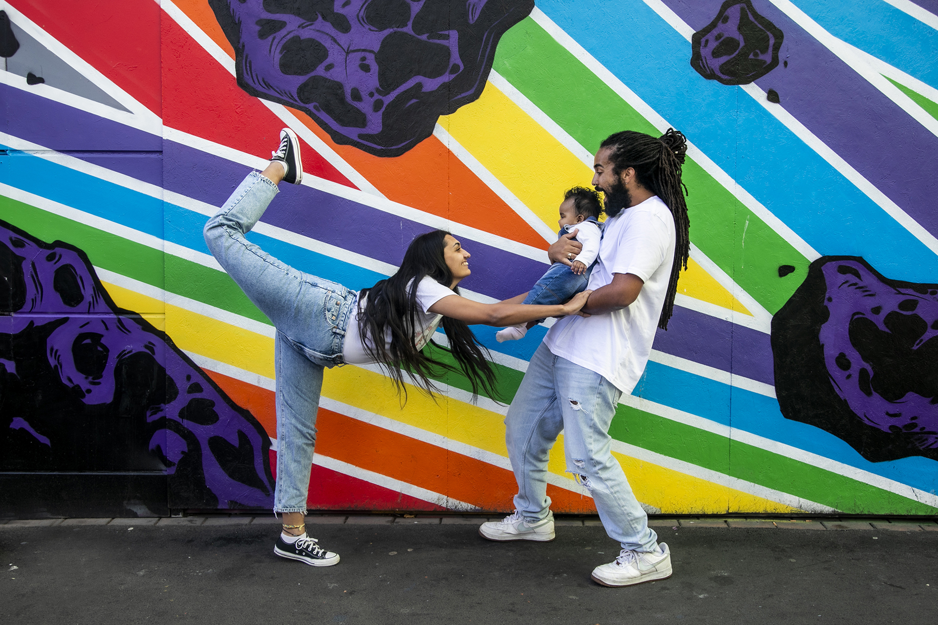 Family of 3. Global majority male with long dreadlocked hair holding baby, global majority female with long black hair, one leg up then reaching our leaning towards the baby. wearing blue jeans, trainers and white tshirts infront of rainbow graffiti wall. Family of 3. Global majority male with long dreadlocked hair holding baby, global majority female with long black hair, one leg up then reaching our leaning towards the baby. wearing blue jeans, trainers and white tshirts infront of rainbow graffiti wall.