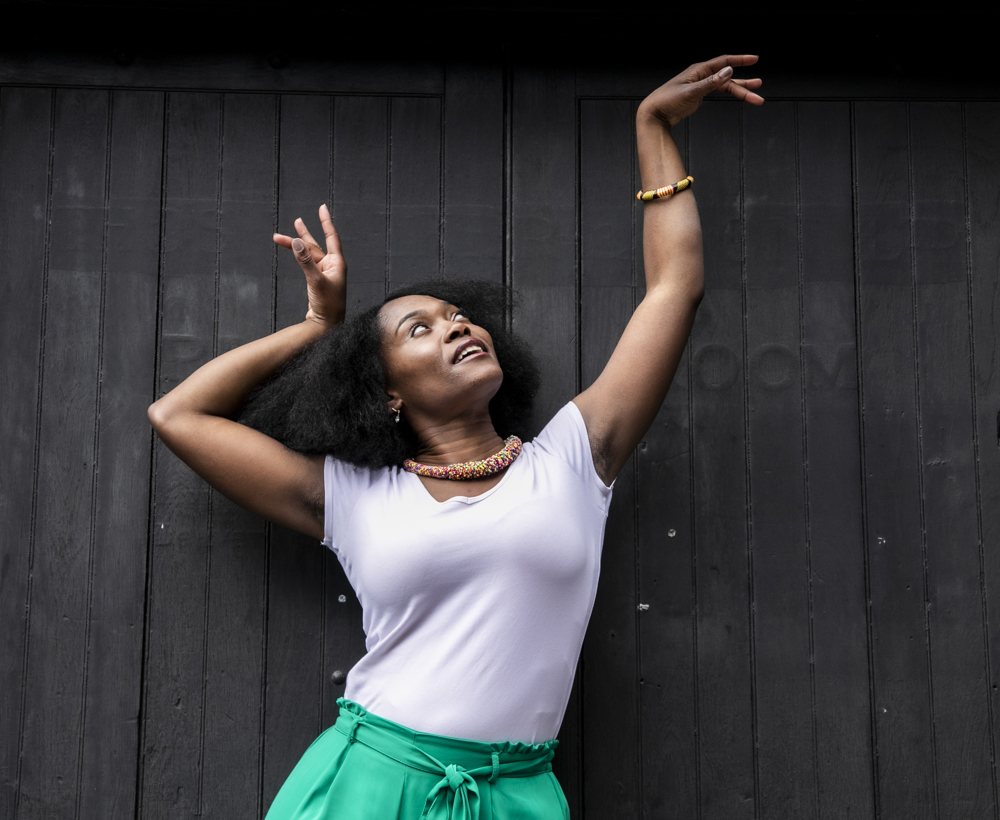 Global majority female dancer with afro, holding one arm out to the top right of the image with eyes looking at a limp wrist and the other hand next to her head. Wearing white top, green trousers and colourful beaded necklace in fornt of black wall