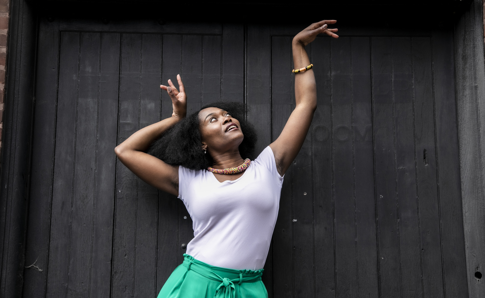 Global majority female dancer with afro, holding one arm out to the top right of the image with eyes looking at a limp wrist and the other hand next to her head. Wearing white top, green trousers and colourful beaded necklace in fornt of black wall Global majority female dancer with afro, holding one arm out to the top right of the image with eyes looking at a limp wrist and the other hand next to her head. Wearing white top, green trousers and colourful beaded necklace in fornt of black wall