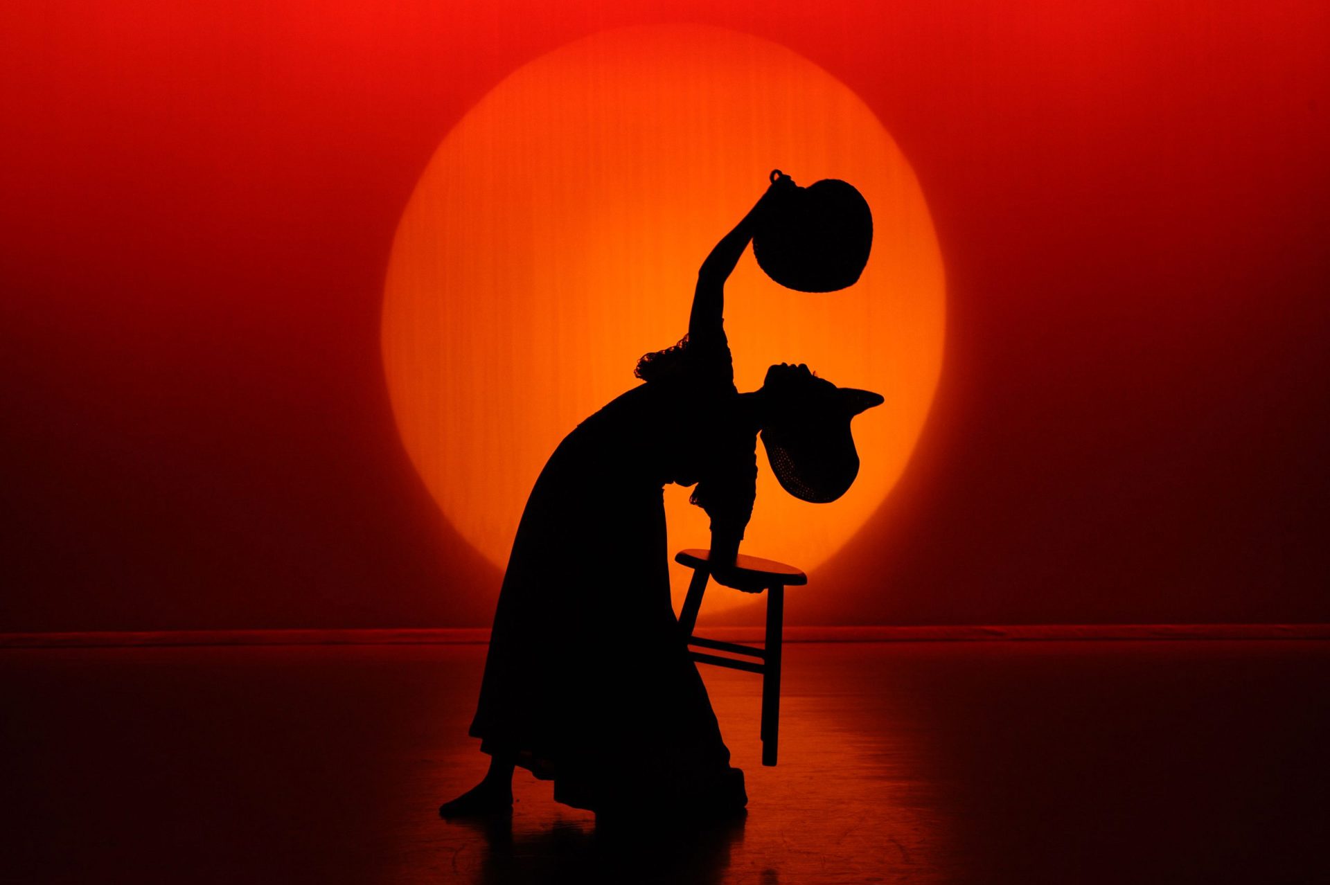 silhouette of dancer doing a backbend holding a tool with one hand and arching over waving a fan to the face wearing a large hat. In front of large orange circle and red background silhouette of dancer doing a backbend holding a tool with one hand and arching over waving a fan to the face wearing a large hat. In front of large orange circle and red background