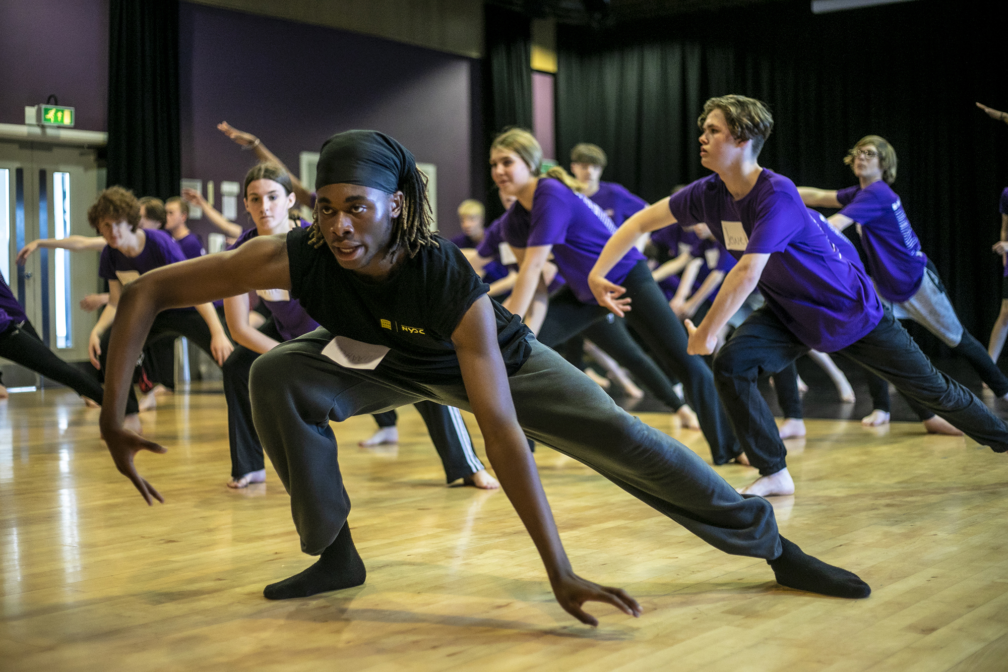 black male dance artist teaching room of young people, teacher and students in deep lunges with one hand touching the floor the other in a wing shape. Teacher wearing black students out of focus wearing purple t-shirts. black male dance artist teaching room of young people, teacher and students in deep lunges with one hand touching the floor the other in a wing shape. Teacher wearing black students out of focus wearing purple t-shirts.