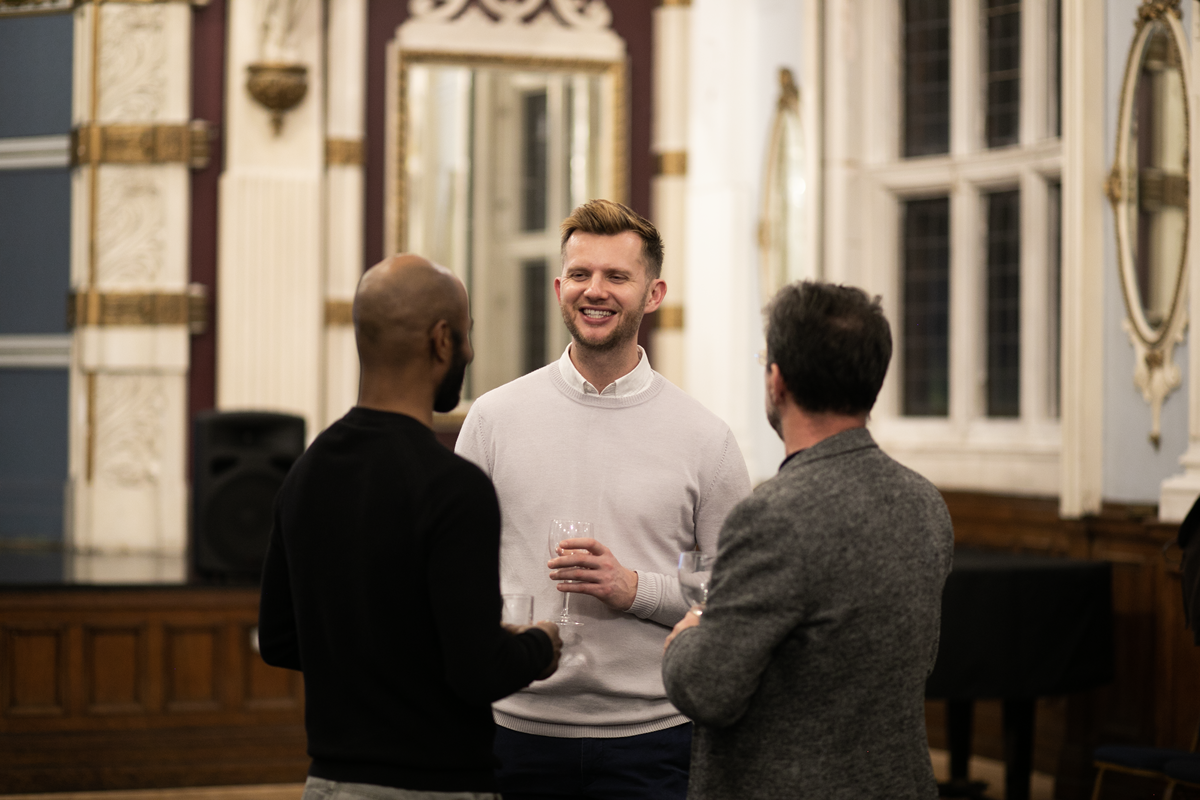 Tall white male in a pale jumper and shirt smiling with a glass of wine whilst talking to two people with their heads to the back of the camera. In a large historial room. 