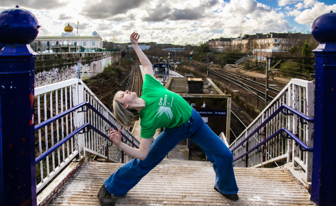 white blonde female lunging with arms high and arched back face pointed to the sky in front of train station platform. Wearing green dance ambassador tshirt and blue jeans