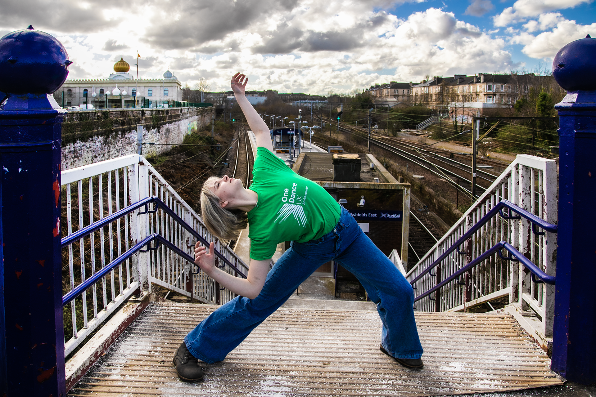 white blonde female lunging with arms high and arched back face pointed to the sky in front of train station platform. Wearing green dance ambassador tshirt and blue jeans