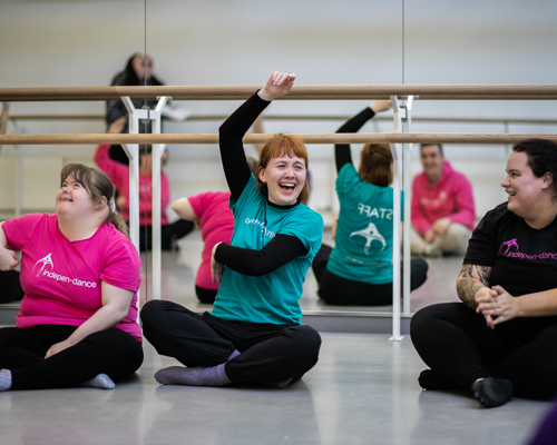 Two white females smiling cross legged, on the left, a disabled dancer in a pink t-shirt, on the right, the group leader raising her arm wearing a teal t-shirt