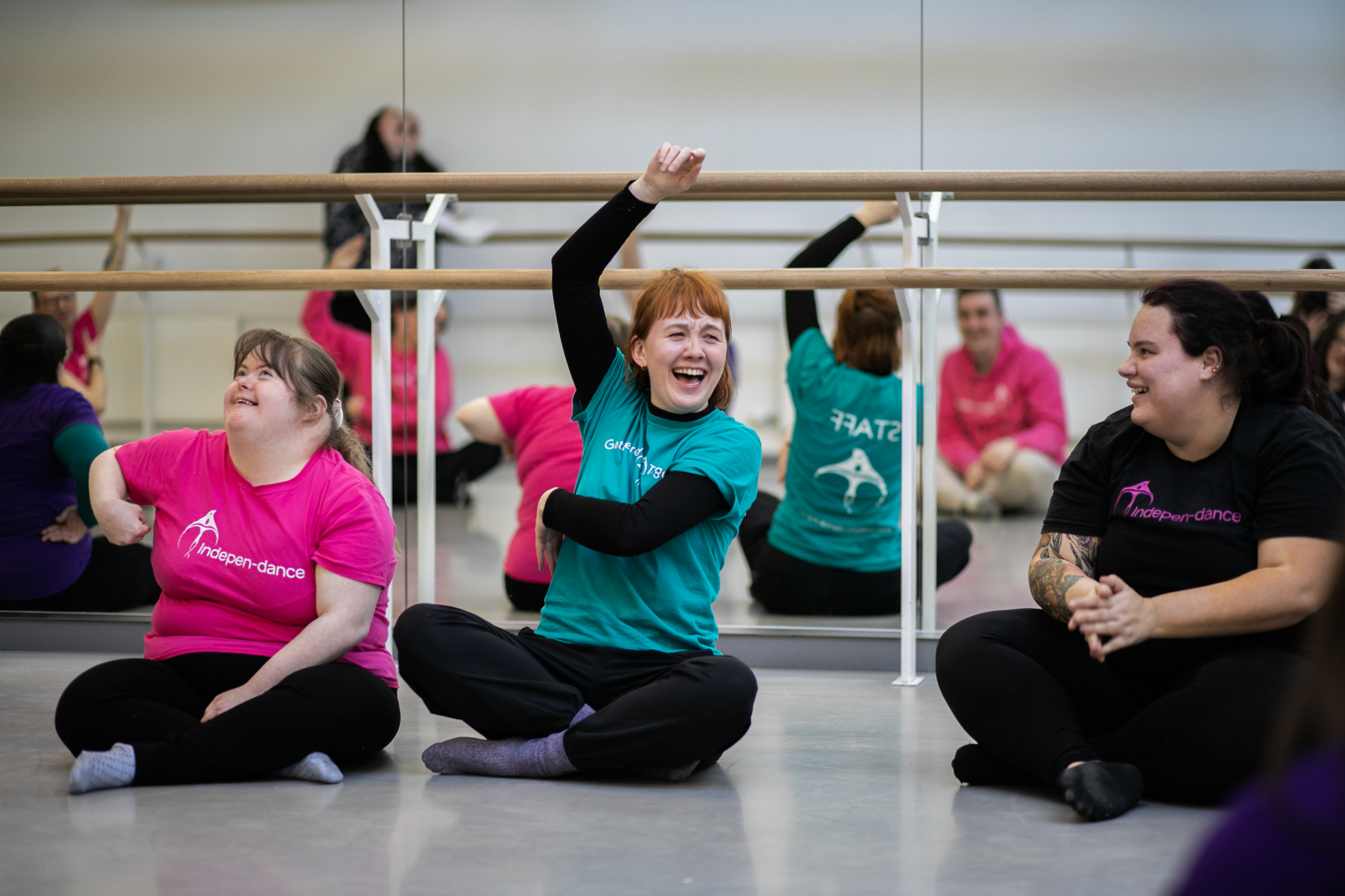 Two white females smiling cross legged, on the left, a disabled dancer in a pink t-shirt, on the right, the group leader raising her arm wearing a teal t-shirt Two white females smiling cross legged, on the left, a disabled dancer in a pink t-shirt, on the right, the group leader raising her arm wearing a teal t-shirt