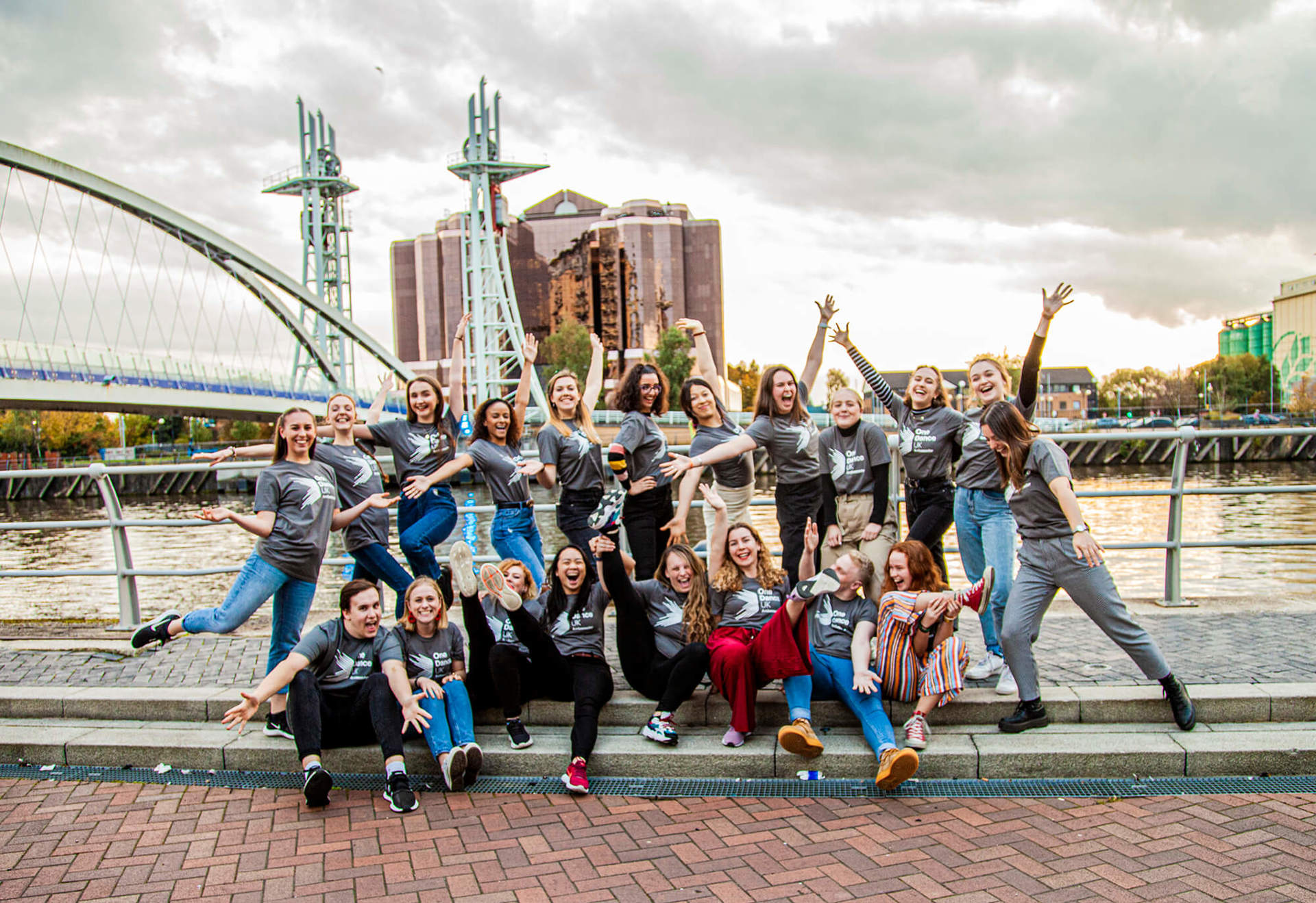 20 male and female dance ambassadors posing in front of Manchester bridge and skyline in the background. Wearing grey Dance ambassador tshirts. 20 male and female dance ambassadors posing in front of Manchester bridge and skyline in the background. Wearing grey Dance ambassador tshirts.
