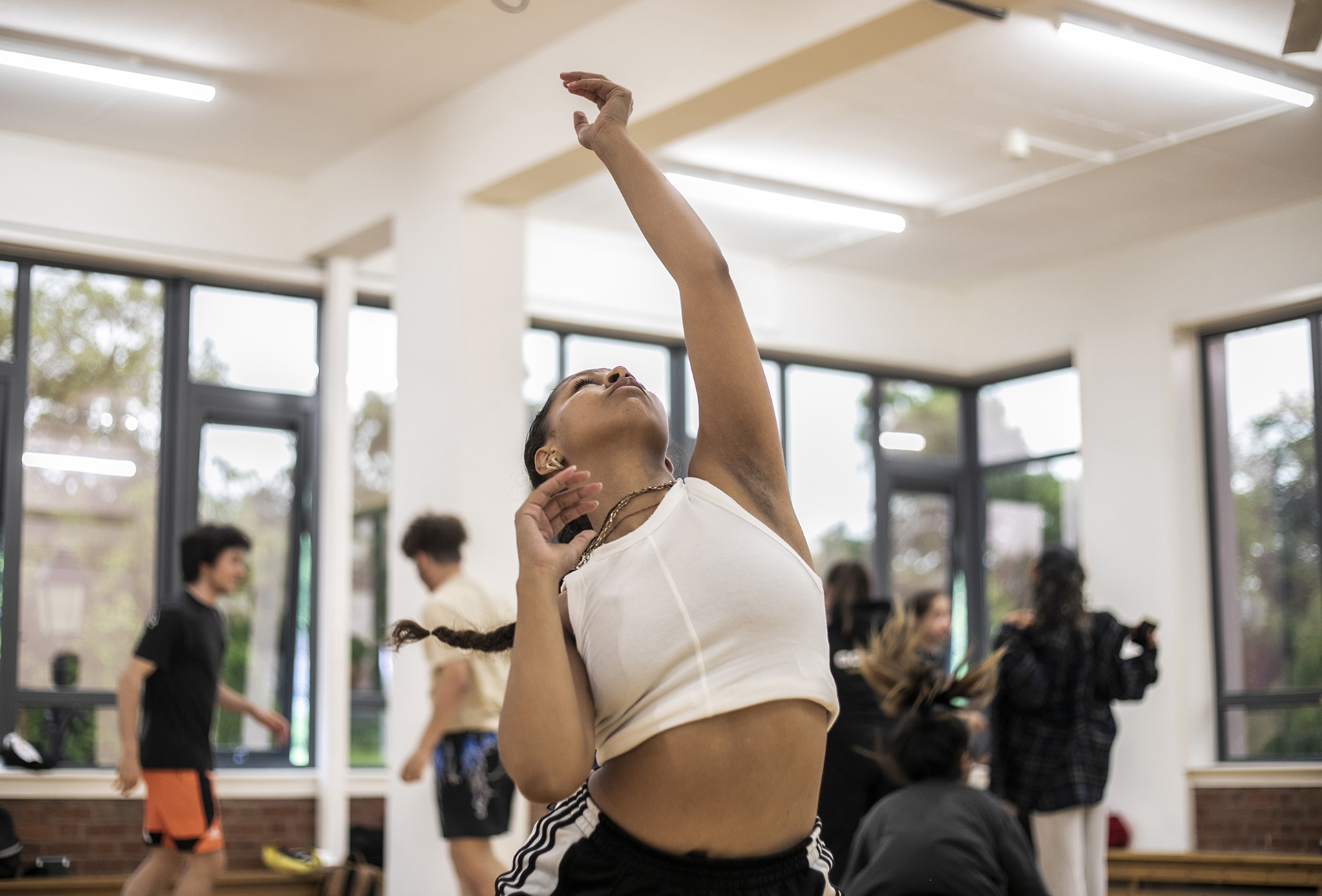 Dancers practising in a dance studio. One global majority female dancer in the centre of the image with one arm straight up looking towards the ceiling, wearing sports wear. 