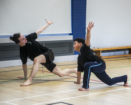 teenage male dance teacher lunging with one arm in the air teaching young boy dancer lunging with the mirrored arm in the air. In sports hall both wearing black PE kit