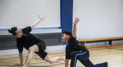 teenage male dance teacher lunging with one arm in the air teaching young boy dancer lunging with the mirrored arm in the air. In sports hall both wearing black PE kit teenage male dance teacher lunging with one arm in the air teaching young boy dancer lunging with the mirrored arm in the air. In sports hall both wearing black PE kit