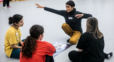 Global majority dance teaching kneeling down with 4 young female dance students gesturing with arms out wide with a mood board in front of them in a dance studio. 