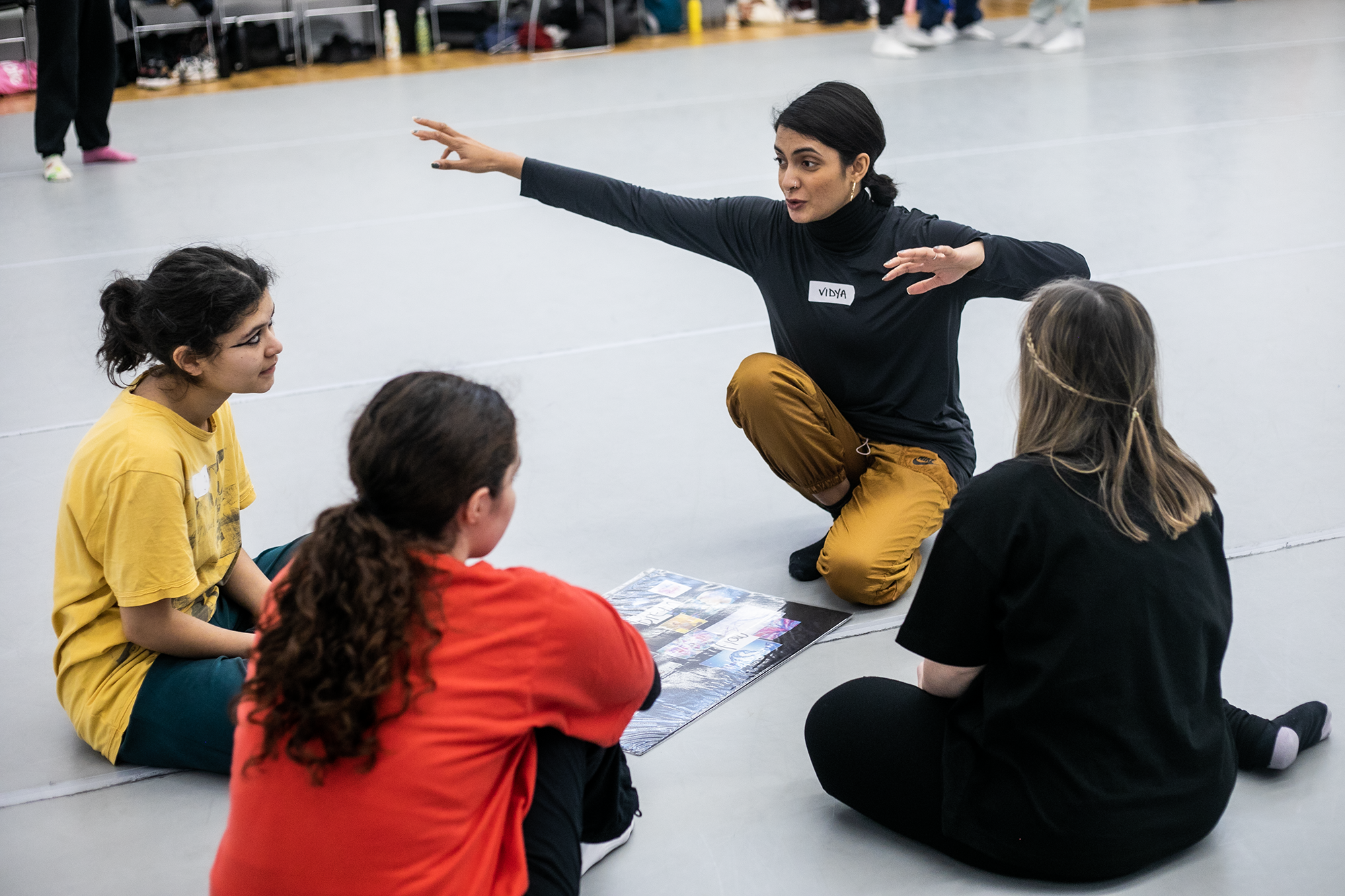 Global majority dance teaching kneeling down with 4 young female dance students gesturing with arms out wide with a mood board in front of them in a dance studio.  Global majority dance teaching kneeling down with 4 young female dance students gesturing with arms out wide with a mood board in front of them in a dance studio.