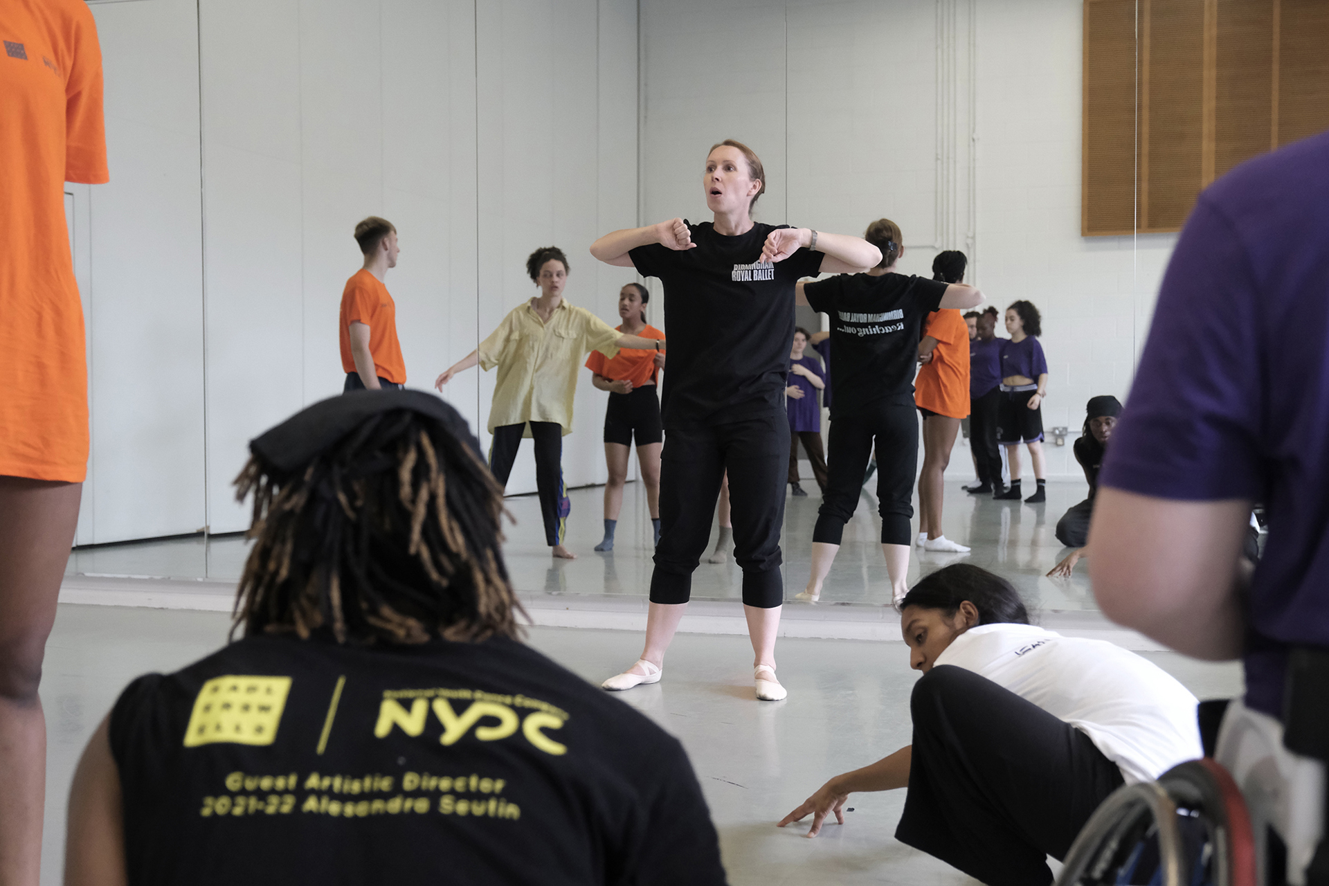 white female dance teacher guestering to a room full of students who are wearing brightly coloured clothes with a mirrored background. In Dance studio.  white female dance teacher guestering to a room full of students who are wearing brightly coloured clothes with a mirrored background. In Dance studio.