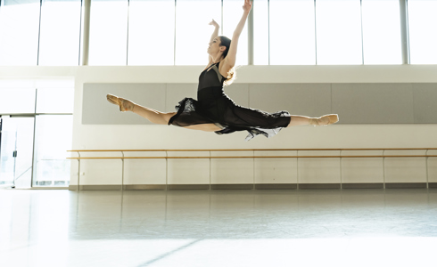 white female dancer in a split leap with arms up wearing a black skirt and leotard. In a dance studio. 