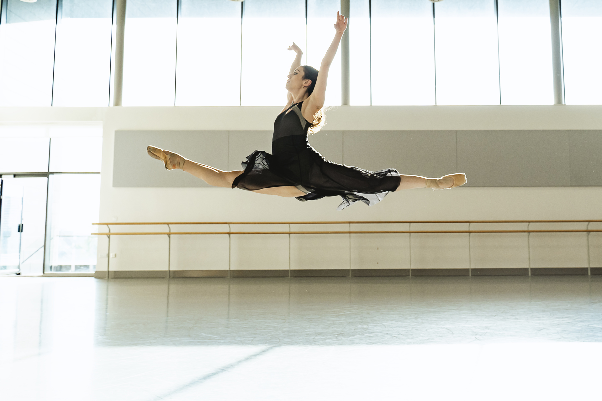 white female dancer in a split leap with arms up wearing a black skirt and leotard. In a dance studio. white female dancer in a split leap with arms up wearing a black skirt and leotard. In a dance studio.