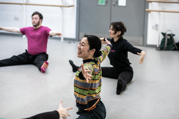 global majority female dancer teaching community group. Arm stretched open sitting with legs wise on the floor in the dance studio. 